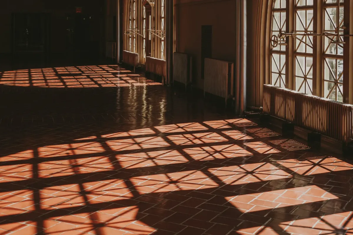 Color photo of the interior of a large hall with a reddish-brown tiled floor and large arched windows casting shadow patterns on the ground.