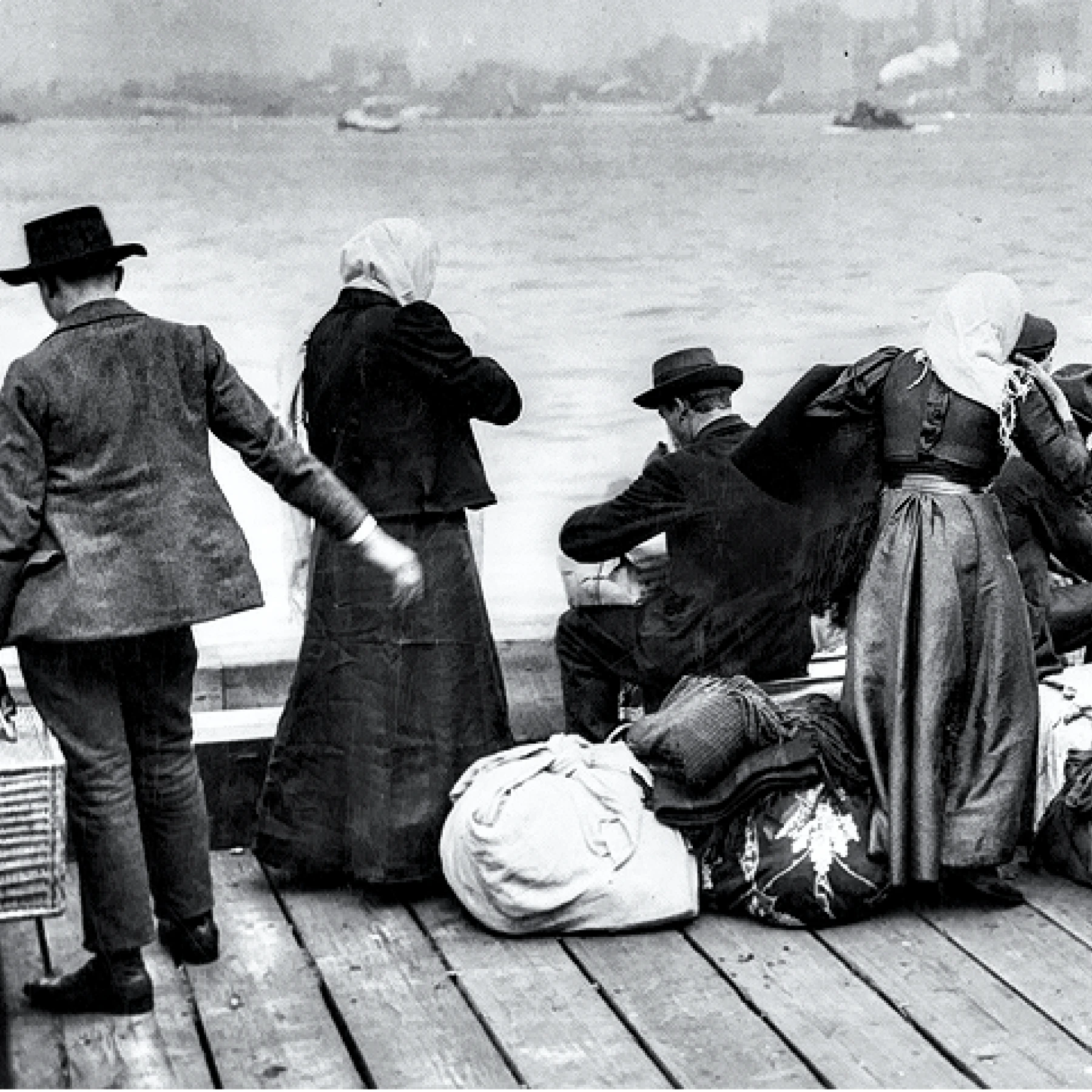 A black and white photo taken from behind a small group of early 20th-century immigrants on a dock with luggage, overlooking a city skyline across the water.