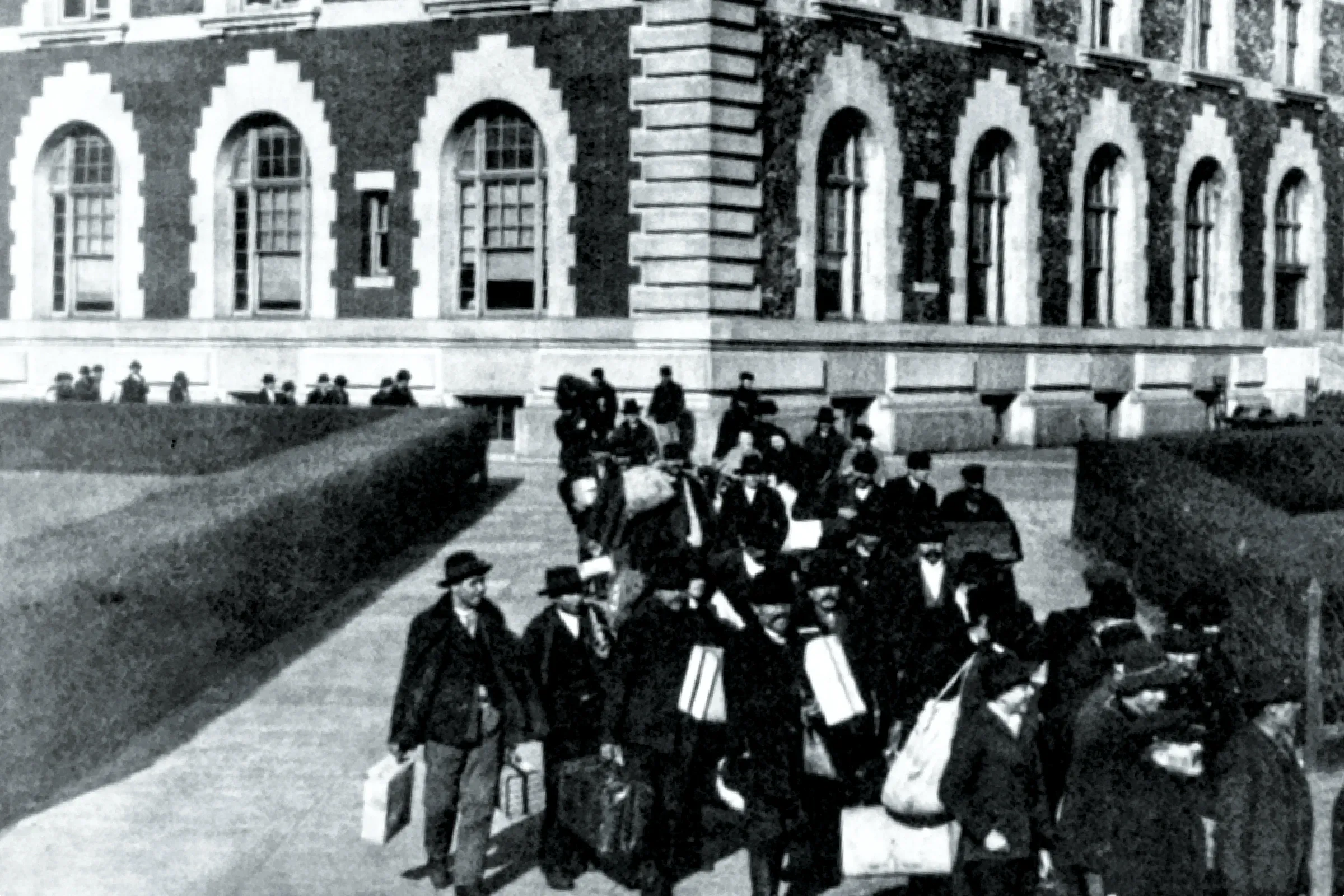 Black and white early 20th-century photo taken above a large group of various immigrants on Ellis Island carrying luggage as they depart from a brick building with arched windows.