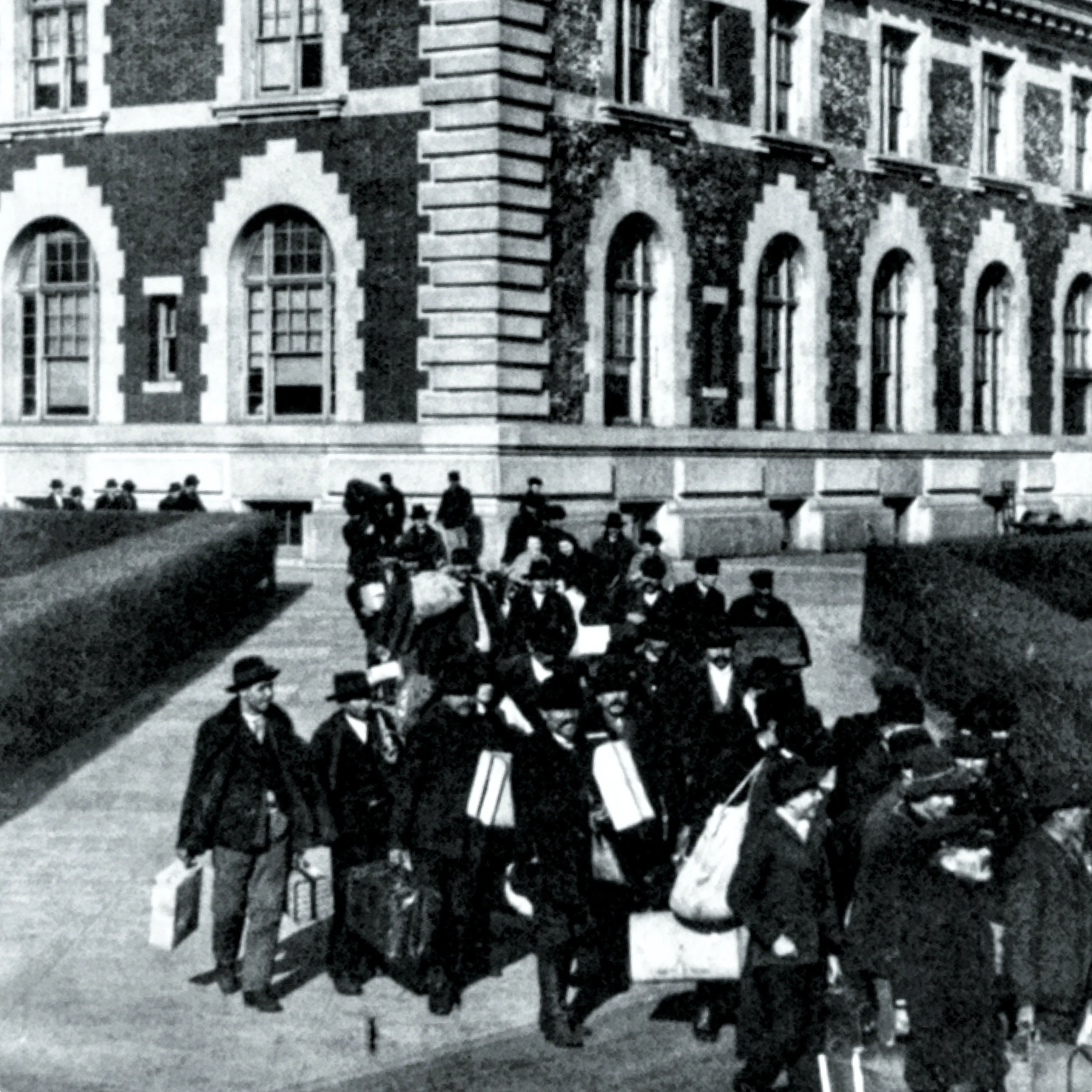 Black and white early 20th-century photo taken above a large group of various immigrants on Ellis Island carrying luggage as they depart from a brick building with arched windows.