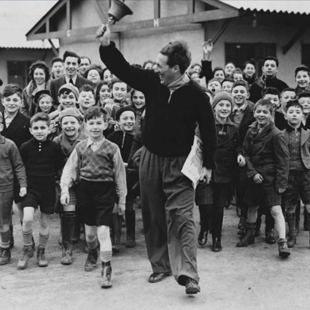 A black and white photo of Harold Stanley joyfully ringing a handheld bell, surrounded by a group of smiling children, in front of a building.