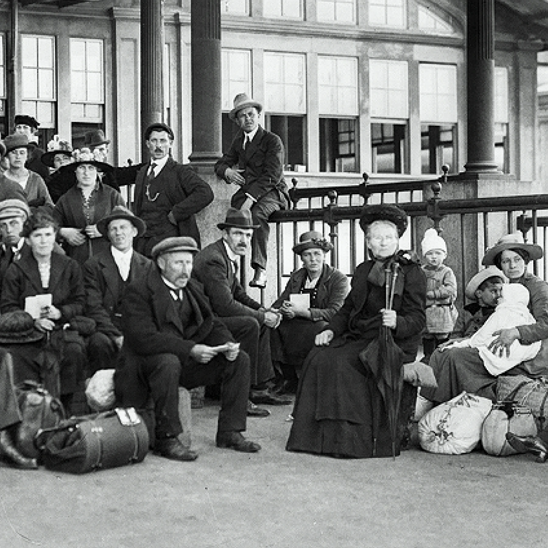 A black and white group photo of early 20th-century light-skinned immigrants of various ages outside a building with their belongings. Some are standing and others are seated. One man sits on the railing.