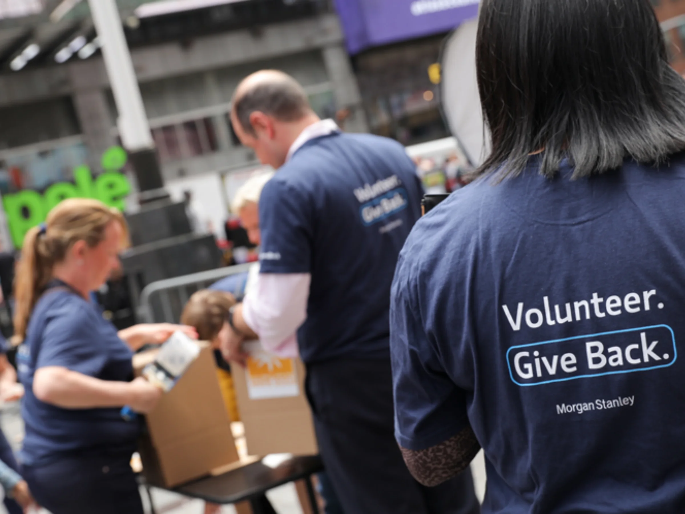 Morgan Stanley volunteers working together at a community event. People handle cardboard boxes and wear navy blue T-shirts that read, “Volunteer. Give Back. Morgan Stanley.”