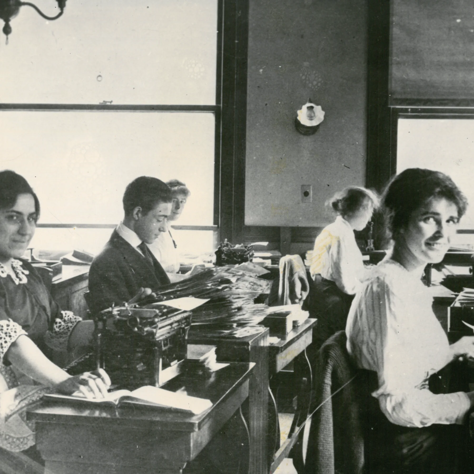 Black and white photo taken in a Liberty Mutual office of employees working at desks with typewriters and stacks of files. One light-skinned woman in the foreground smiles at the camera.