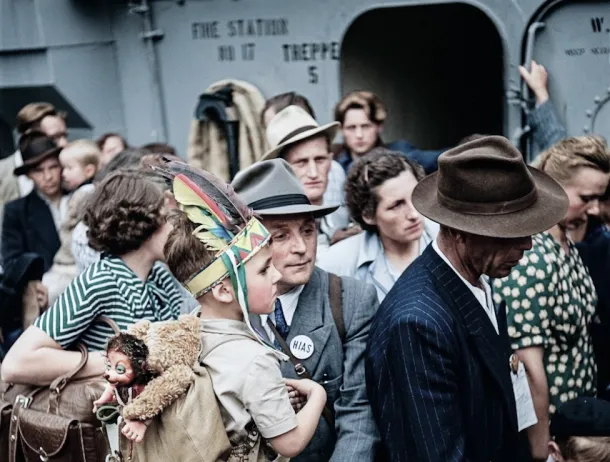 Close-up photo of light-skinned men, women, and children on the deck of a ship. A man in the front wears a grey suit, a fedora, and a button reading "HIAS”. He carries a boy wearing a colorful, feathered, Native American-style headdress, and backpack with a teddy bear.
