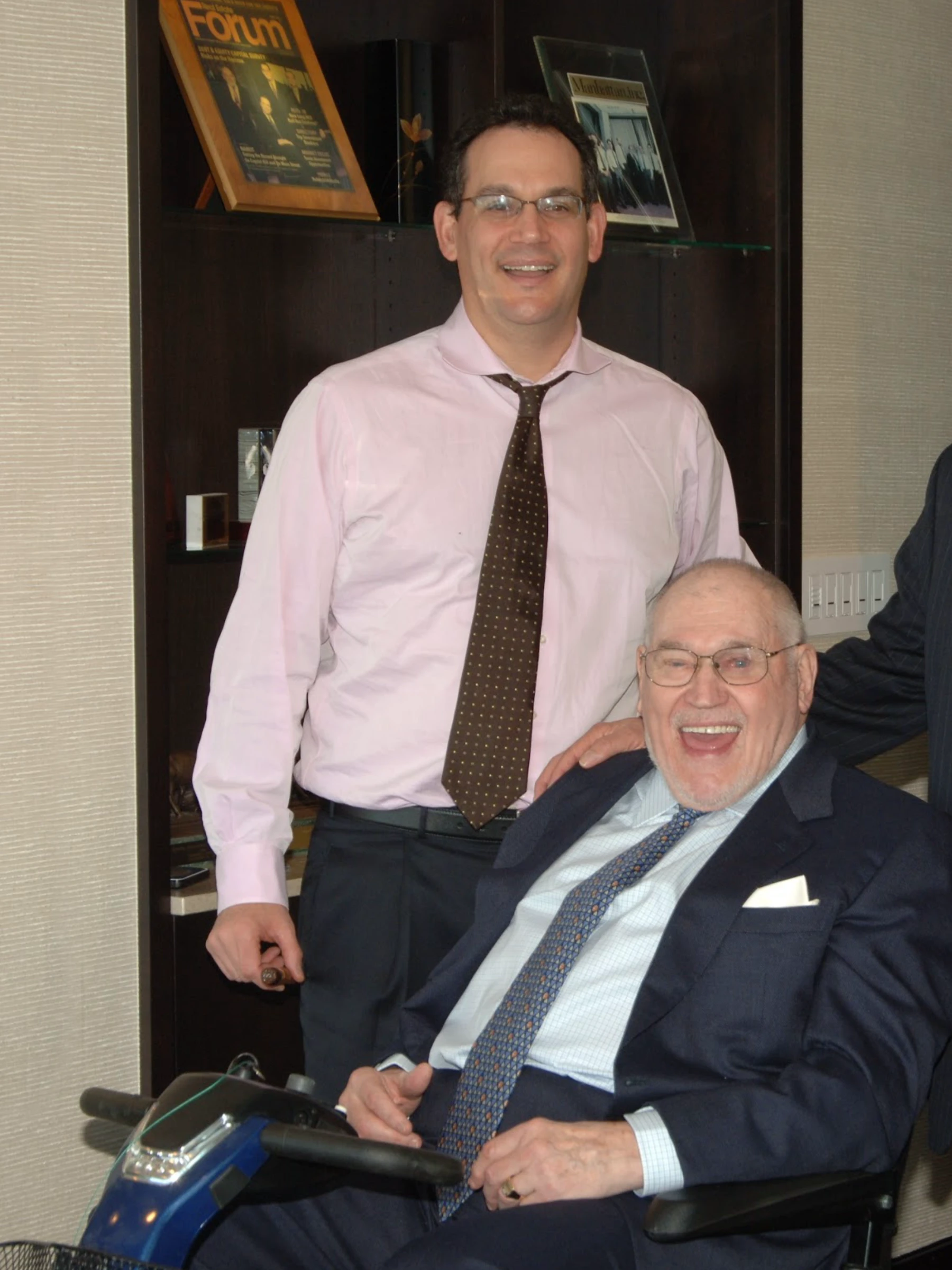Color photo of a light-skinned, senior gentleman wearing a suit and tie sitting in a motorized wheelchair. A light-skinned, younger man in a pink dress shirt and dark tie stands behind him. Both smile widely at the camera.