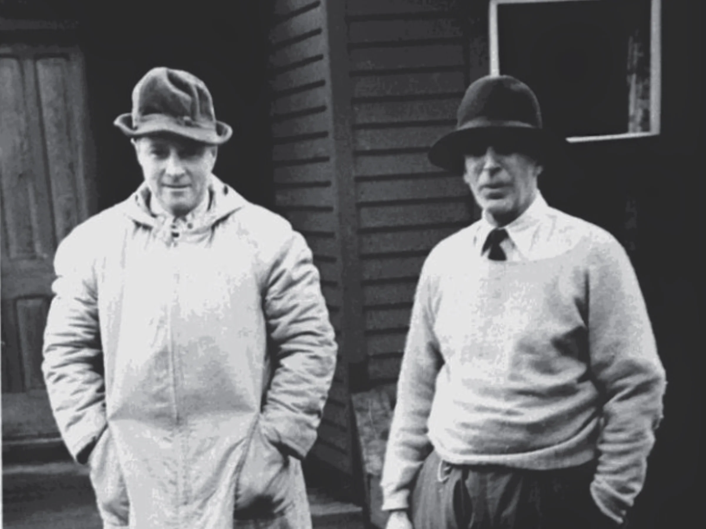 A black and white photo of two men standing outdoors in front of a wooden building. Stanley, left, wears a light coat and misshapen hat, while Morgan wears a sweater and felt hat.