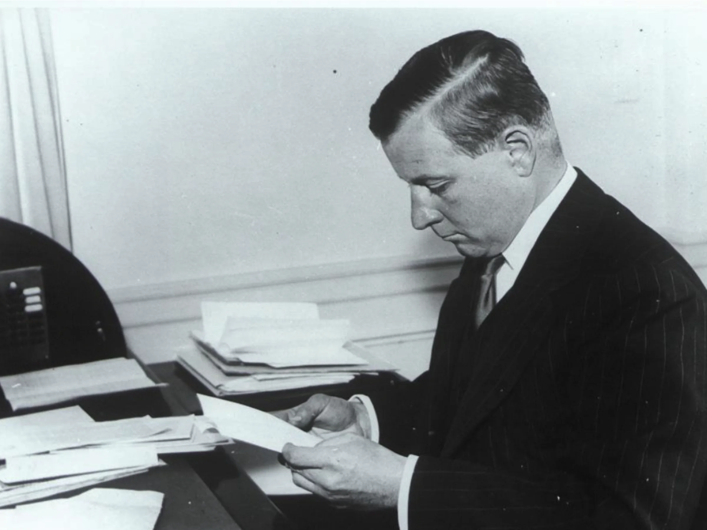 A black and white side profile of a light-skinned man in a pinstripe suit reading a piece of paper while seated at a desk cluttered with documents.