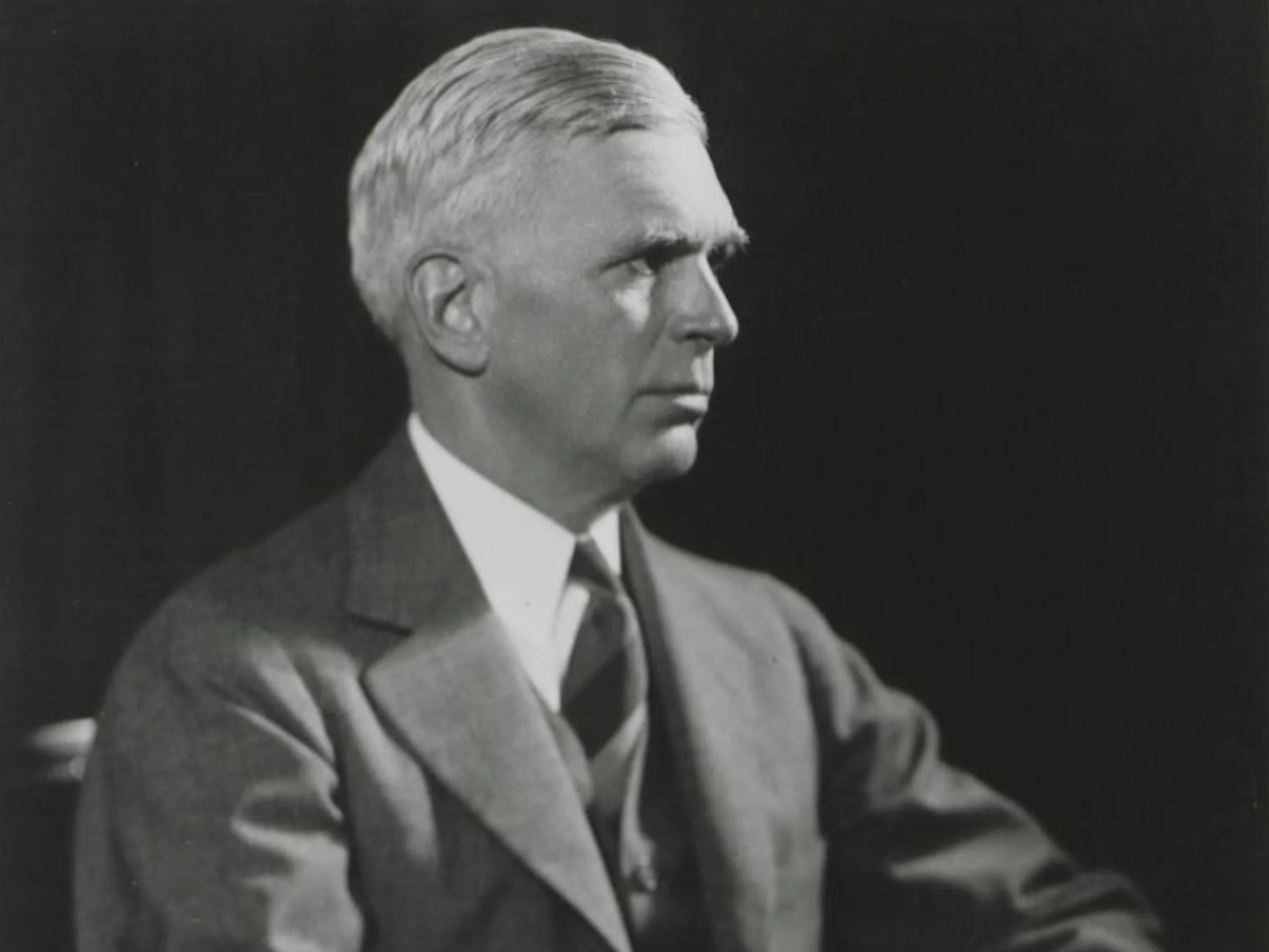 A black and white photo of the side profile of a light-skinned, older man with light-colored hair in a formal suit, set against a dark background.
