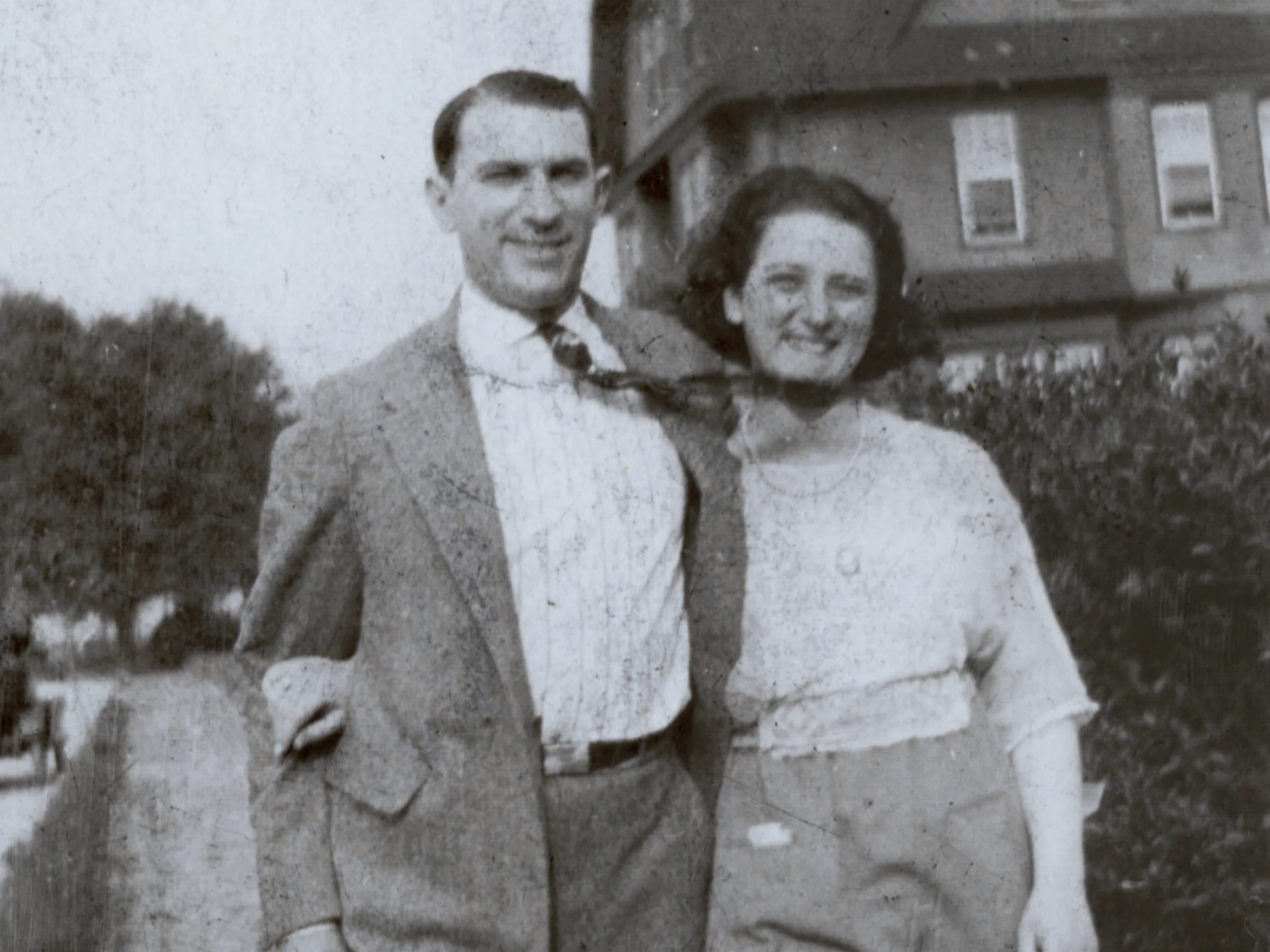 A black and white photo of a smiling couple standing together on a sidewalk. The man wears a suit and the woman wears a blouse and trousers, with shrubbery and a building behind them.