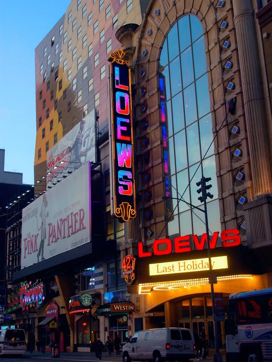 A color photo of the exterior of a Loews Theater with illuminated signage, including a marquee for the film "Last Holiday," set against a clear blue sky.