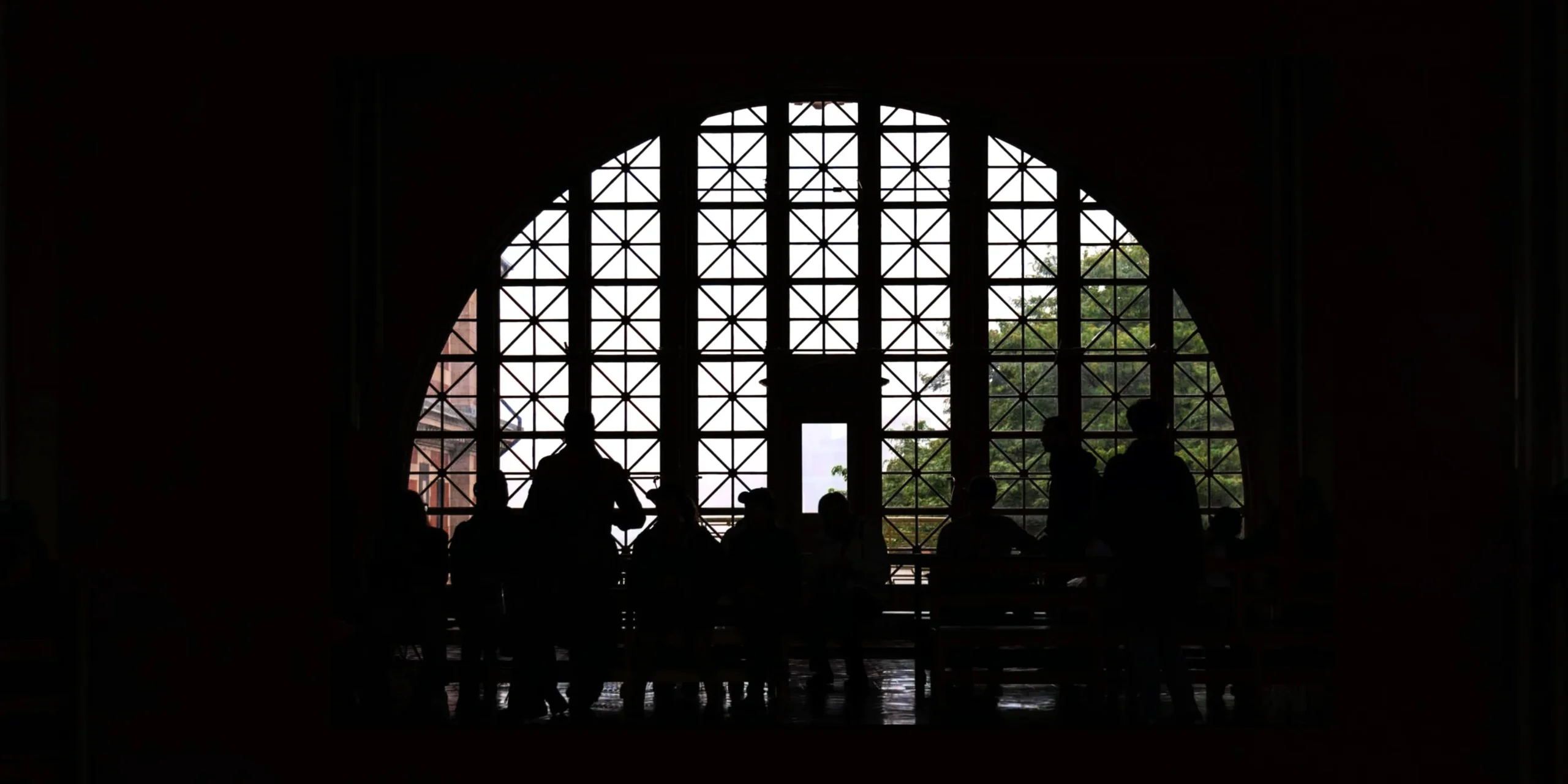 Silhouetted figures standing indoors, looking toward a large, grid-patterned, semi-circular window. Bright daylight shines through the glass, highlighting trees outside and contrasting sharply with the dark, shadowed interior.