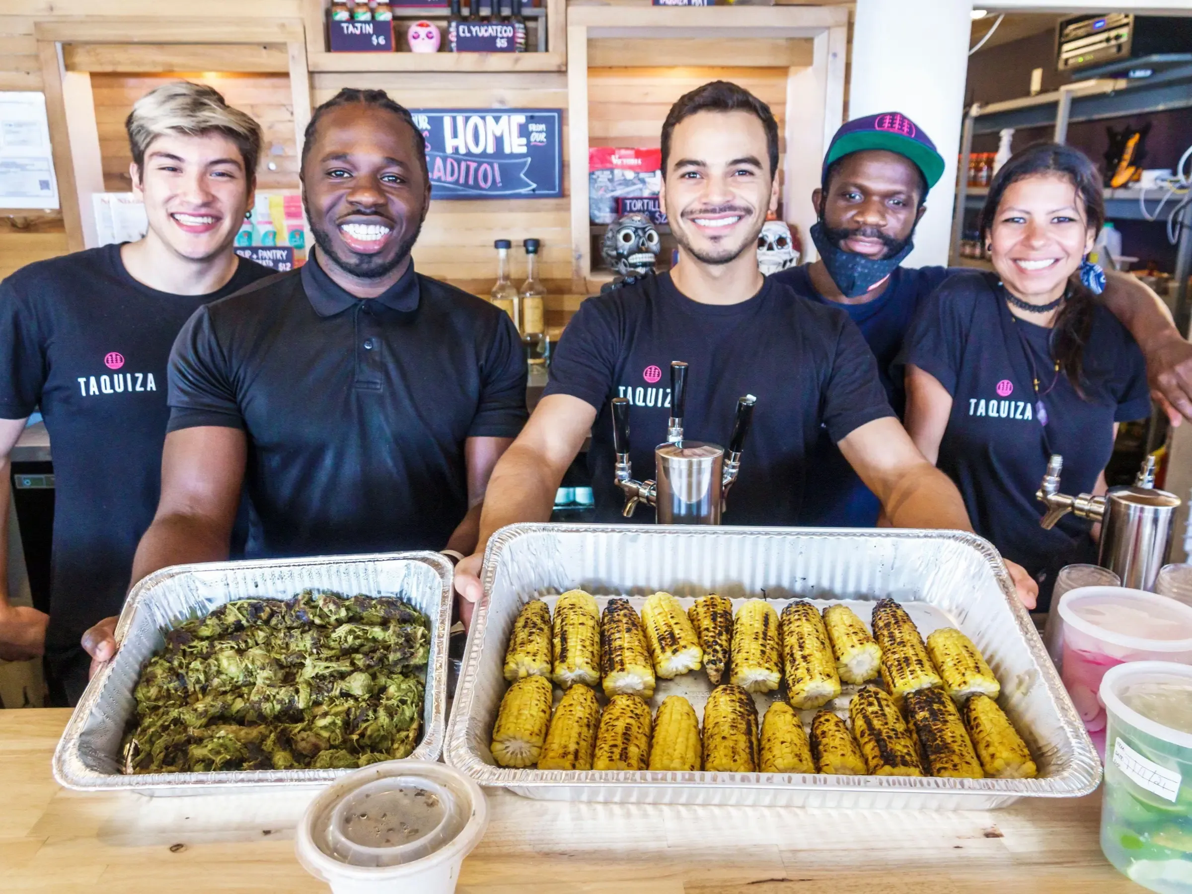 A modern group photo in a food service setting. Five smiling employees of various skin tones in dark, TAQUIZA-branded shirts stand behind a counter holding two food trays, one with grilled corn and another with sautéed greens.