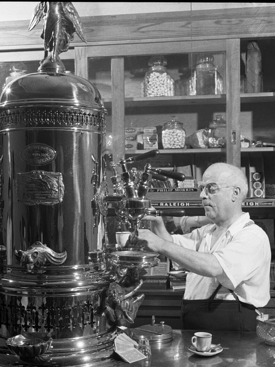 Black and white early 20th century photo of a white man in a short-sleeved white shirt using a large, ornate espresso machine with a decorative figure on top. Jars filled with various items line wooden shelves behind him.