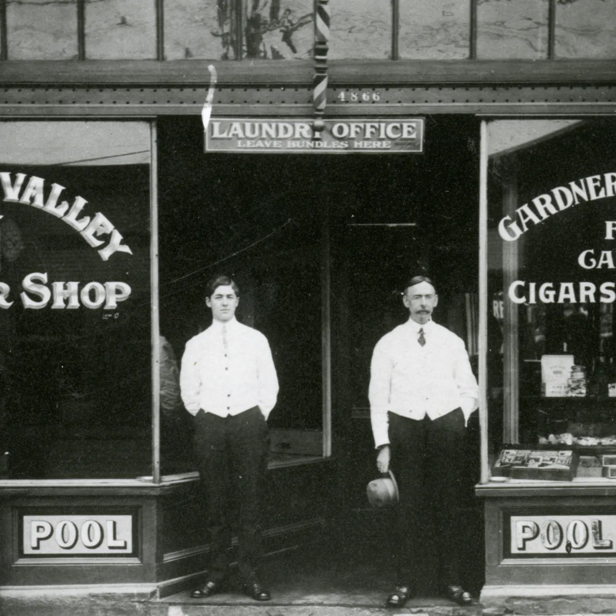 Black and white early 20th century photo of two men in white vests and black pants standing in the doorway of a storefront. Barbershop poles are at left and above them a sign reads “Laundry Office”.