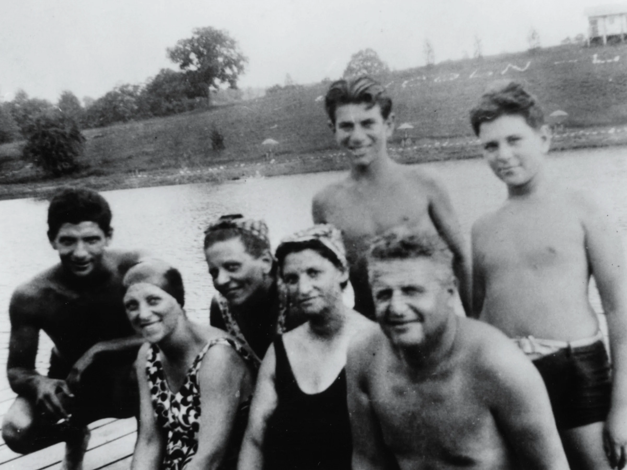 A black and white photo of seven people of wide age ranges in swimwear at a summer camp by a lake, posing together, with trees and a grassy slope in the background.