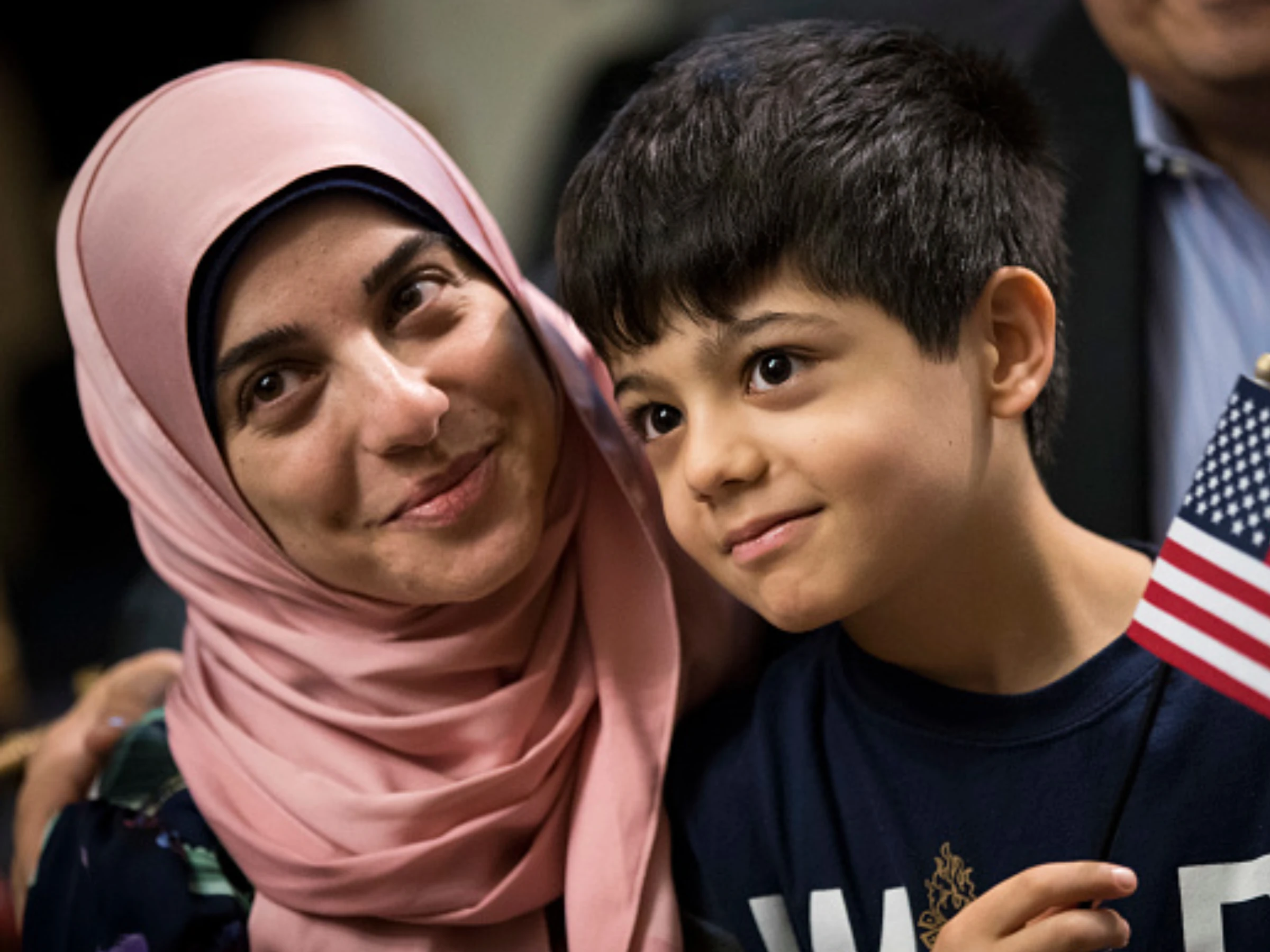 A close-up photo of a woman and child both with medium-brown skin. She wears a pink hijab, and the child holds a small American flag. Both are smiling.