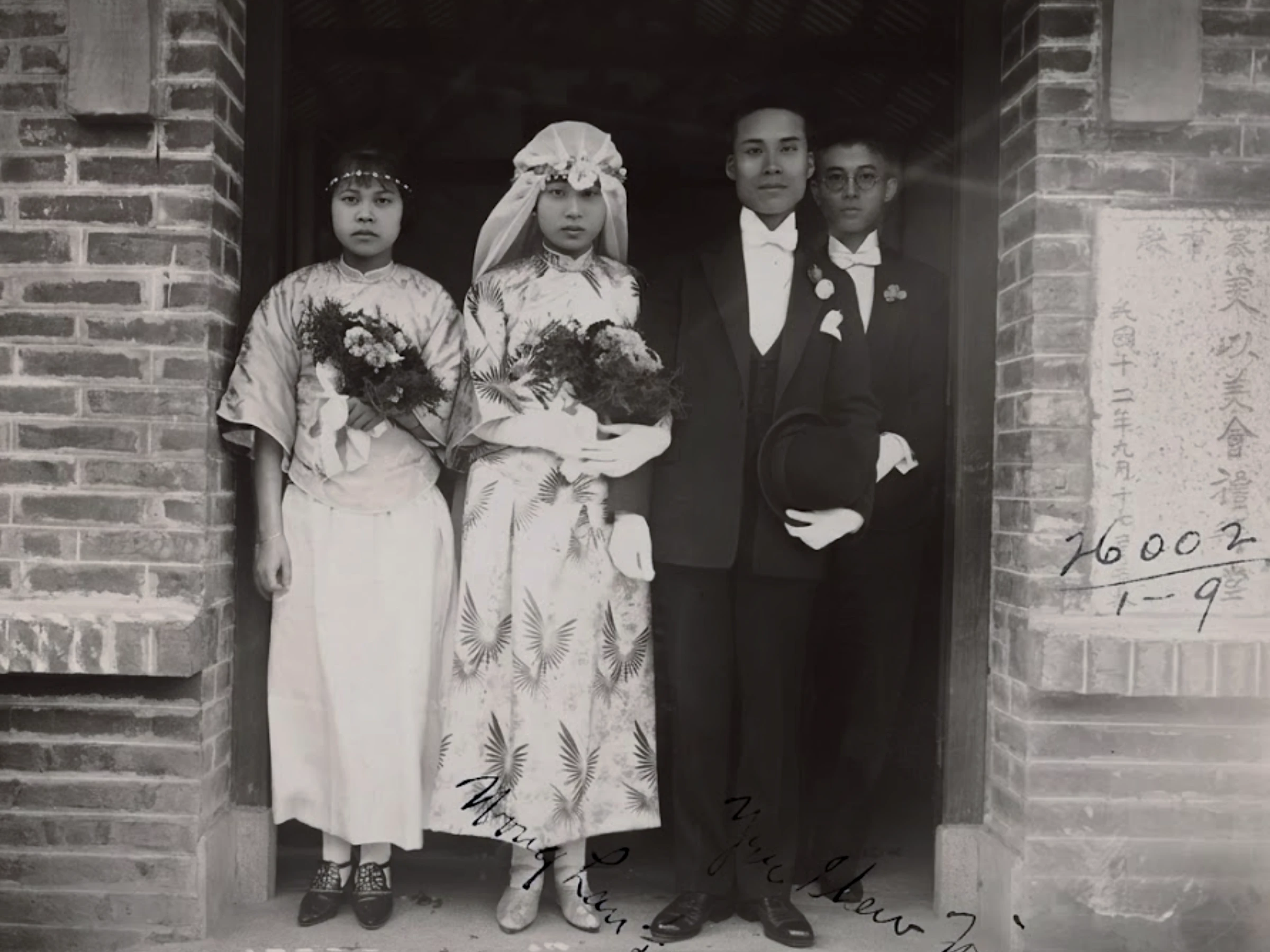 A black and white photo of four people at a Chinese wedding. Two women in elegant dresses on the left hold bouquets, while two men in formalwear stand on their right. One man holds a top hat, and another man is partially visible in the background.