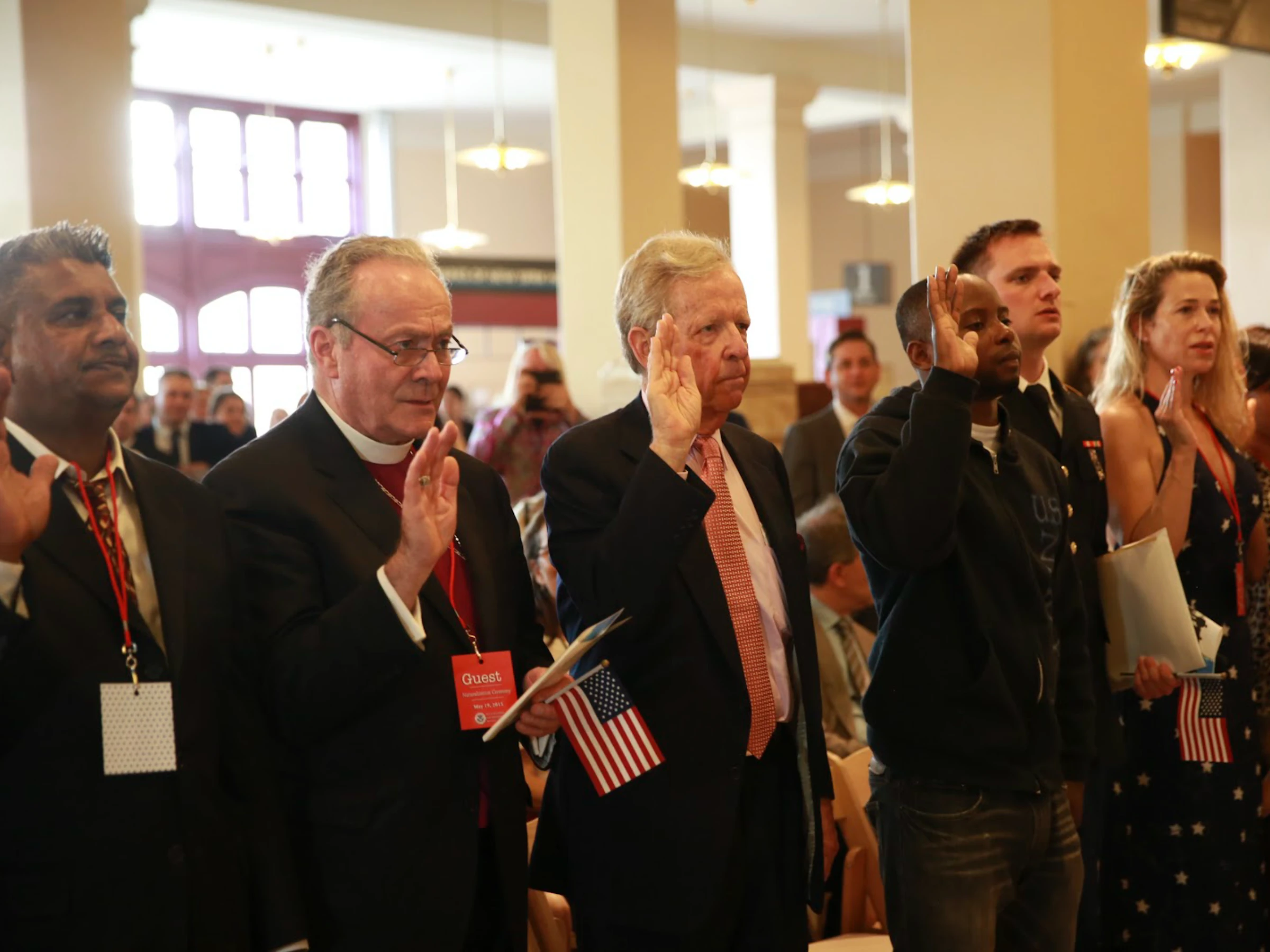 A group of men and women with varying skin tones stand in an open room with columns. Each person has their right hand raised, participating in a naturalization ceremony to become U.S. citizens.