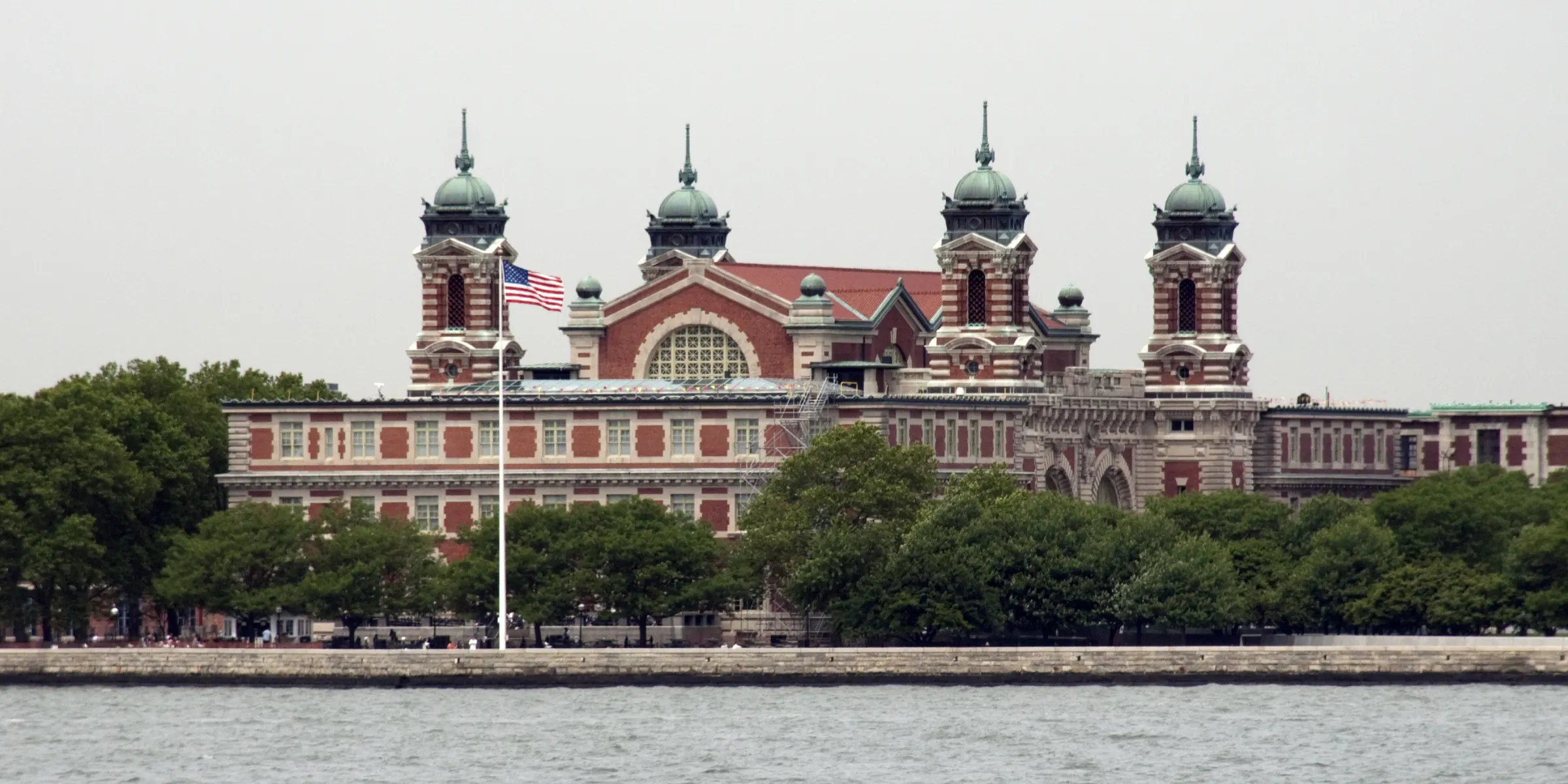 Ellis Island photographed from a distance, featuring red brick architecture with green domes and spires. Trees surround the building, a U.S. flag is in the foreground, with the waterfront in the bottom of the image.