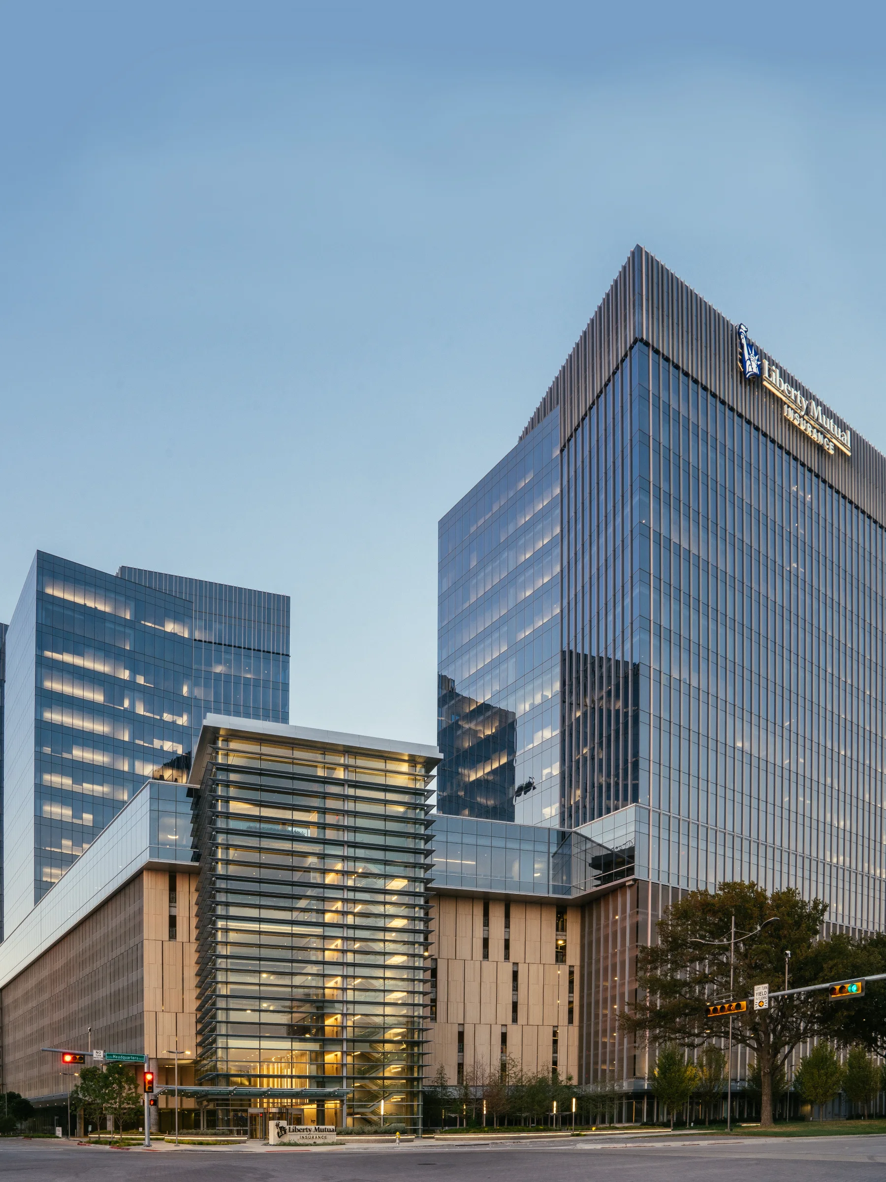 A color photo of a cluster of modern skyscrapers on a sunny day with glass facades in Plano, Texas. The Liberty Mutual logo is visible on the rightmost building.