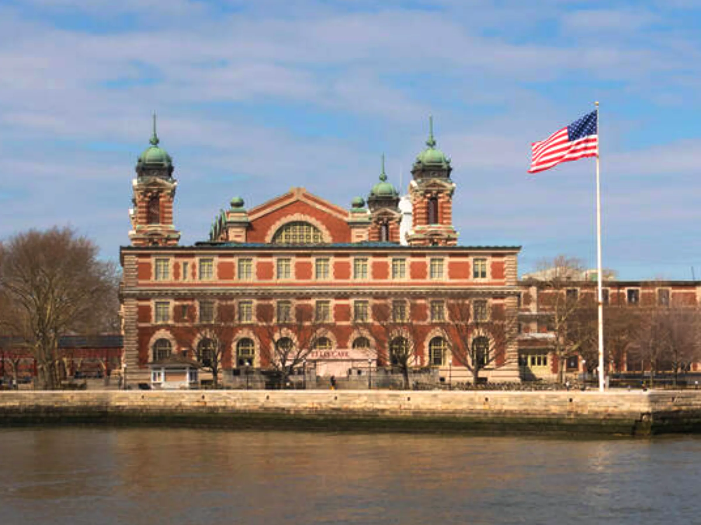 Ellis Island Immigration Museum, a red brick building with green domes, flanked by trees and a flagpole displaying the American flag, set against a blue sky.