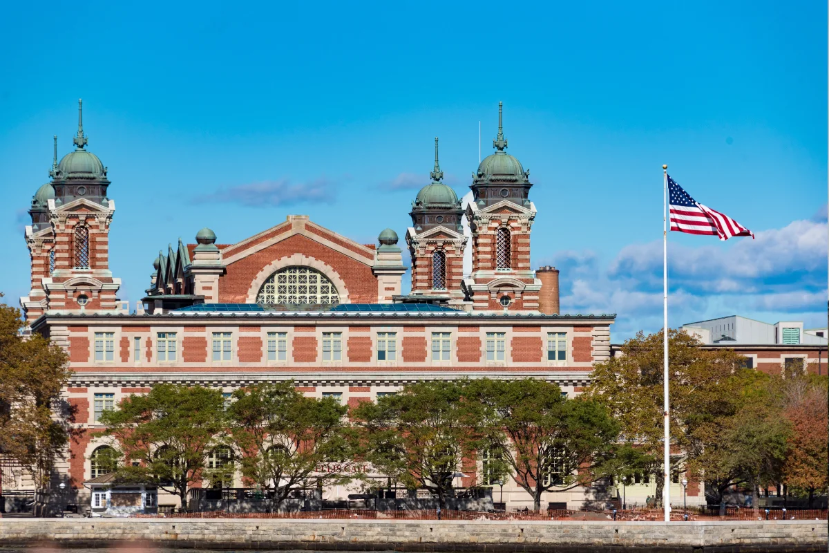 The Ellis Island Museum’s Main Building on a sunny day with copper-domed towers, trees, and an American flag on a flagpole in front.