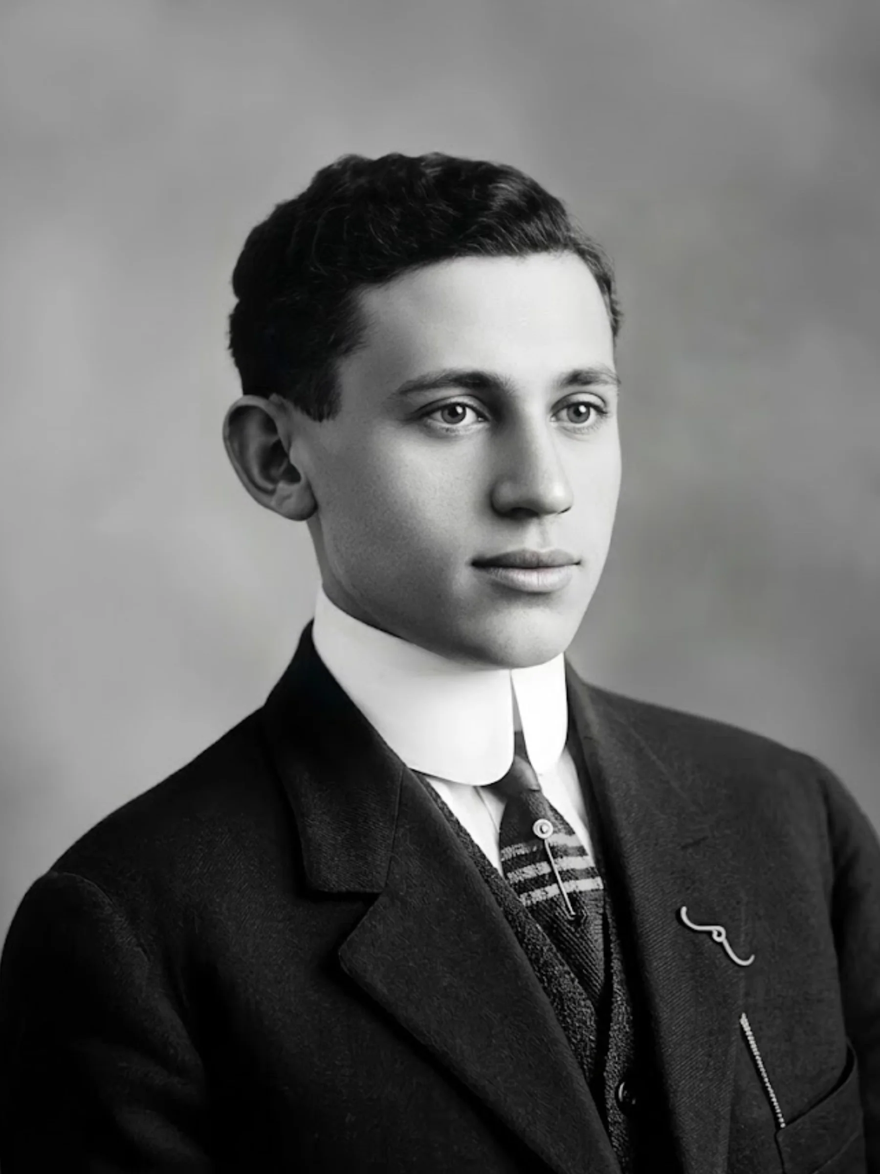 Black and white early 20th-century portrait of a young man against a gray background. He wears a black suit jacket, a white high-collared dress shirt, and a tie