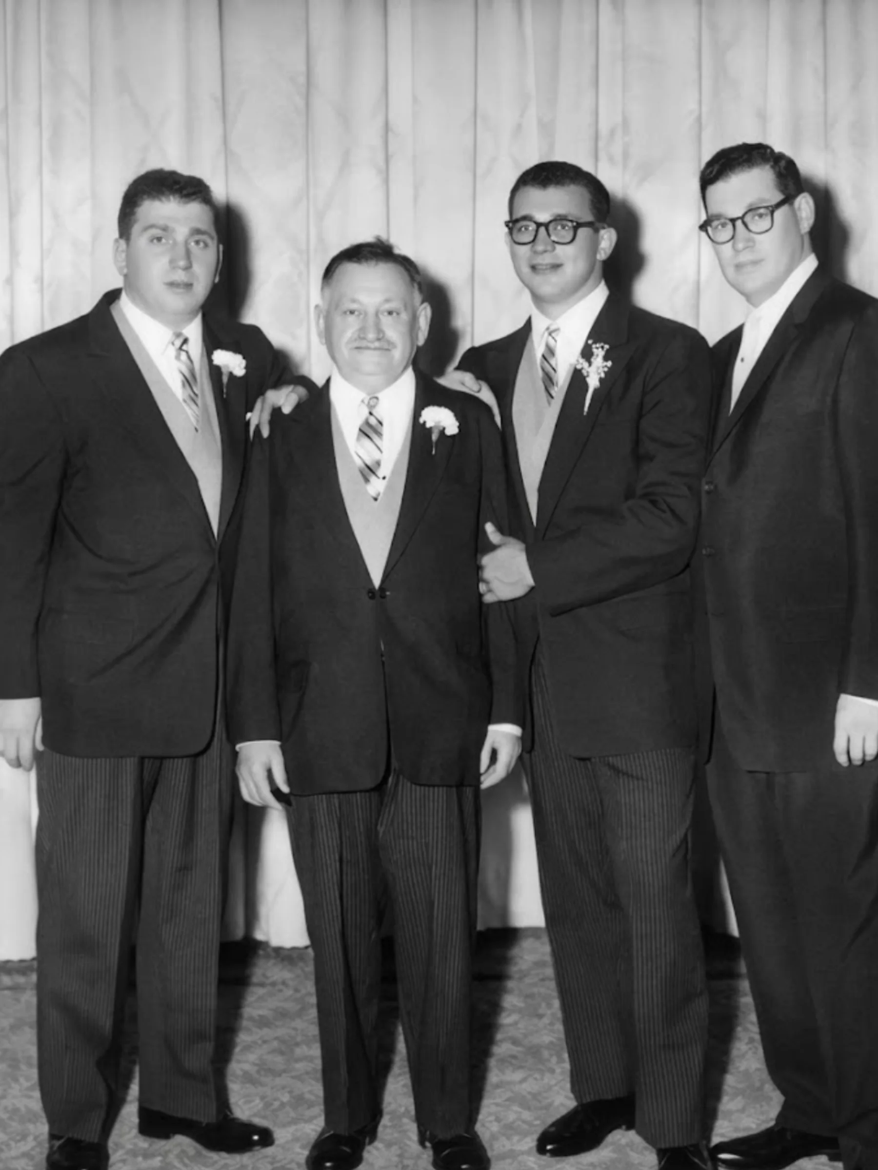 Black and white mid-20th-century photo of four light-skinned men in black suits with white flowers on their lapels, posing together against a light backdrop.