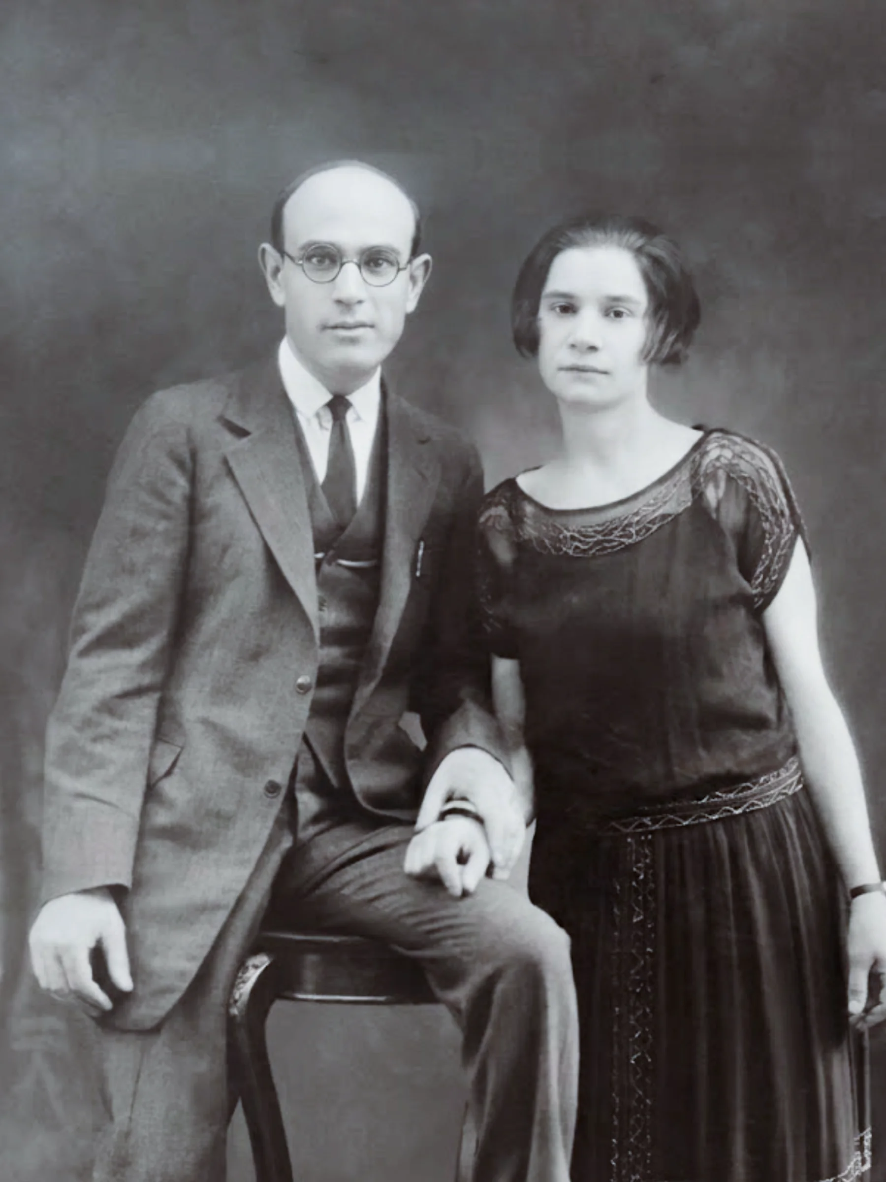 Black and white portrait of a light-skinned man and woman holding hands. He is seated, wearing glasses and a 3-piece suit. She stands, wearing a dark dress with short sleeves.