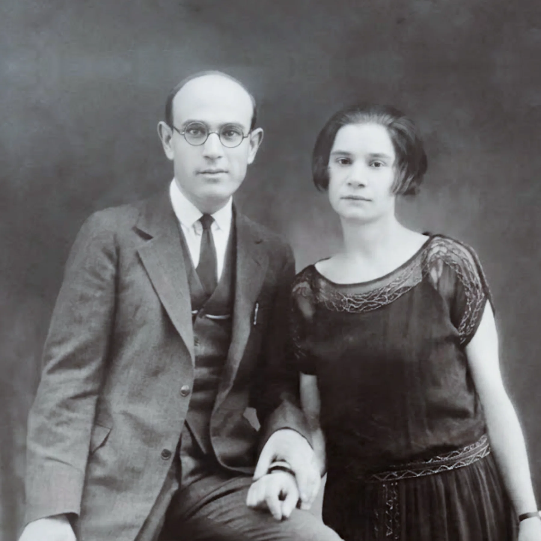 Black and white portrait of a light-skinned man and woman holding hands. He is seated, wearing glasses and a 3-piece suit. She stands, wearing a dark dress with short sleeves.