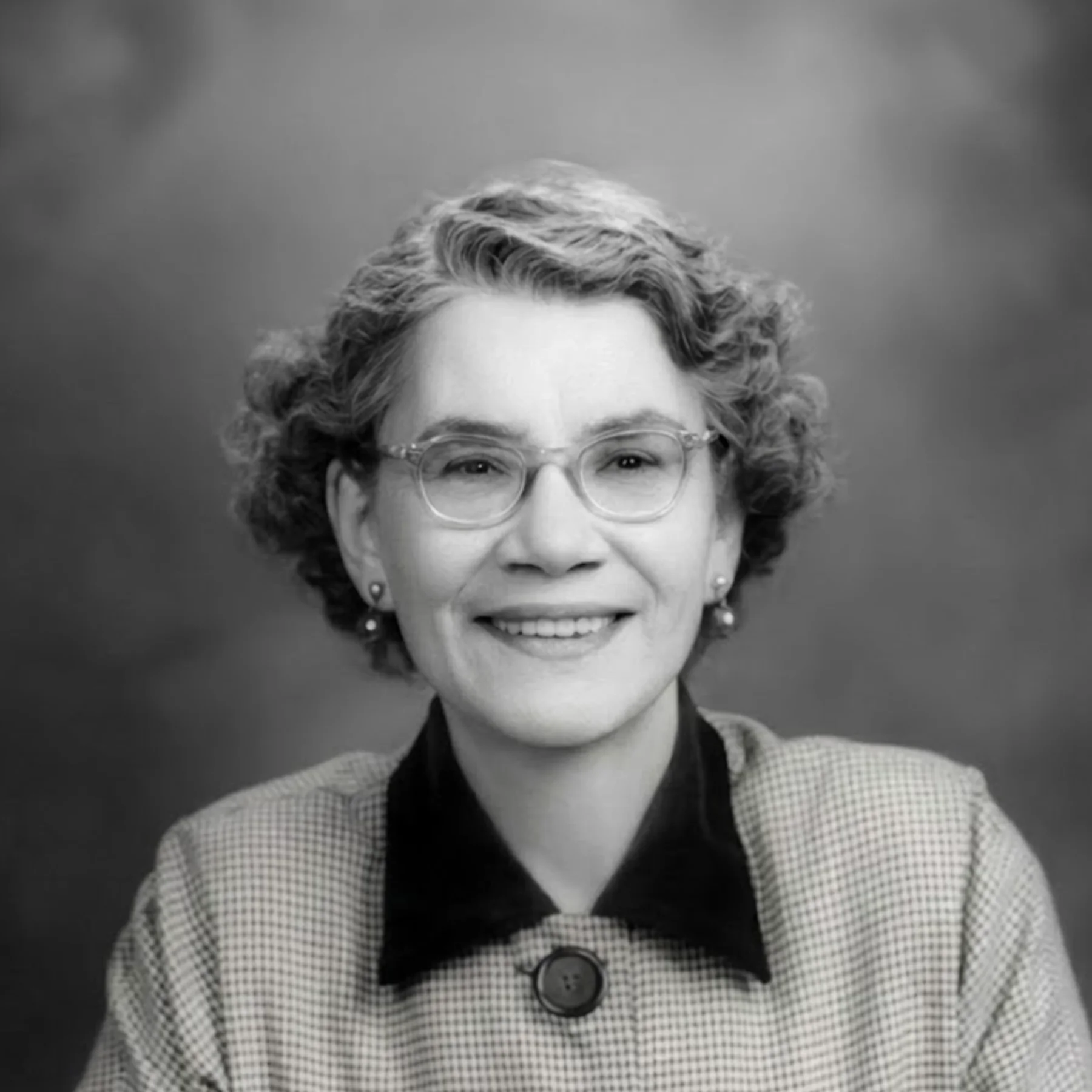 Black and white portrait of a smiling, light-skinned woman’s face. She has short, curly hair, wears wire-rimmed glasses, earrings, and a houndstooth top with a black collar.