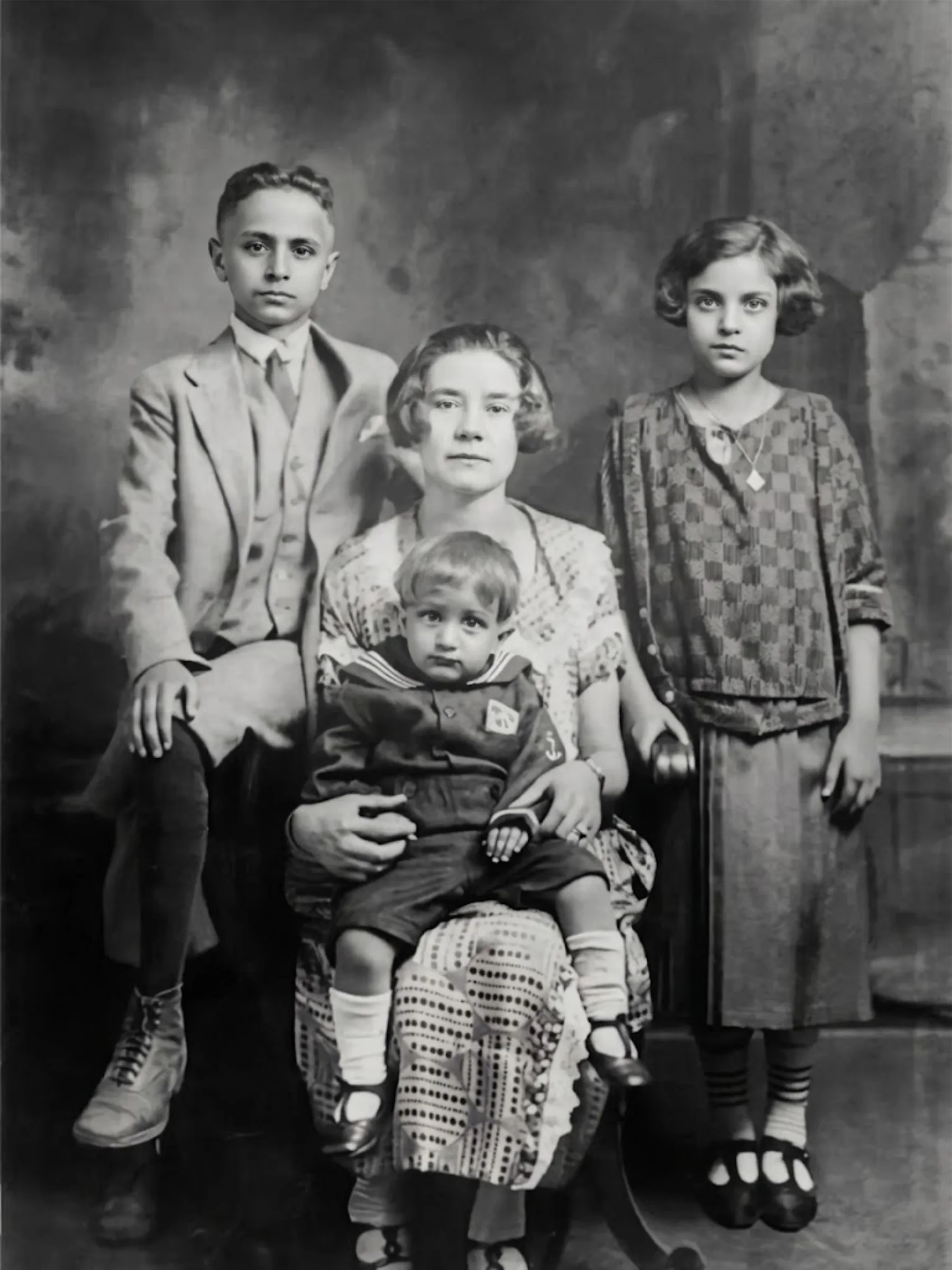 Black and white portrait photo of a light-skinned family featuring a seated woman with a child on her lap. A girl in a dress and a boy in a suit stand on either side of her.