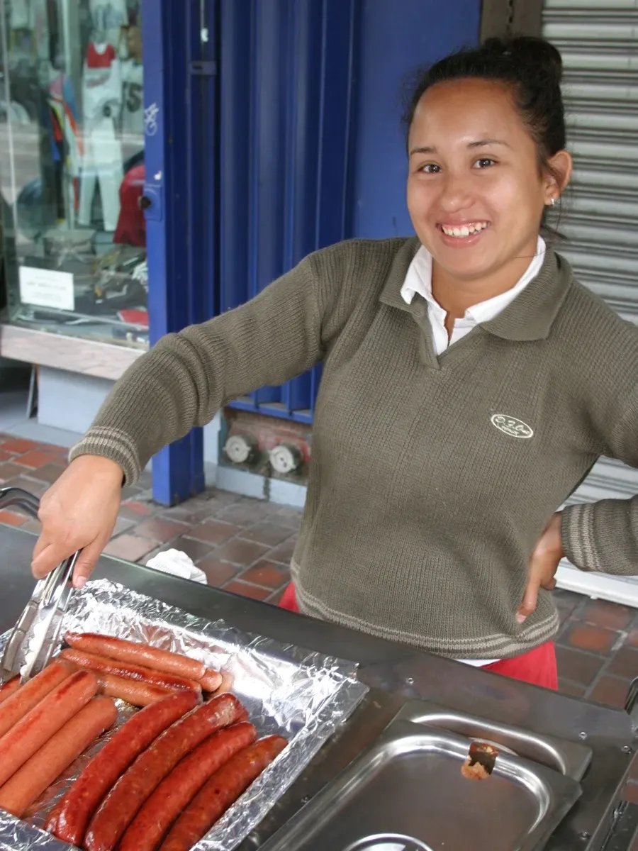 A woman with medium-brown skin in a brown long-sleeved shirt smiling while using tongs to turn sausages in a foil-lined pan.