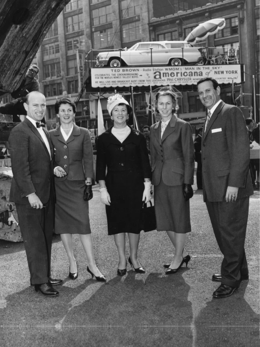 A black and white photo of three women and two men, all light-skinned, posing together in formal attire outdoors, with a vintage Chrysler car and large advertisement in the background.
