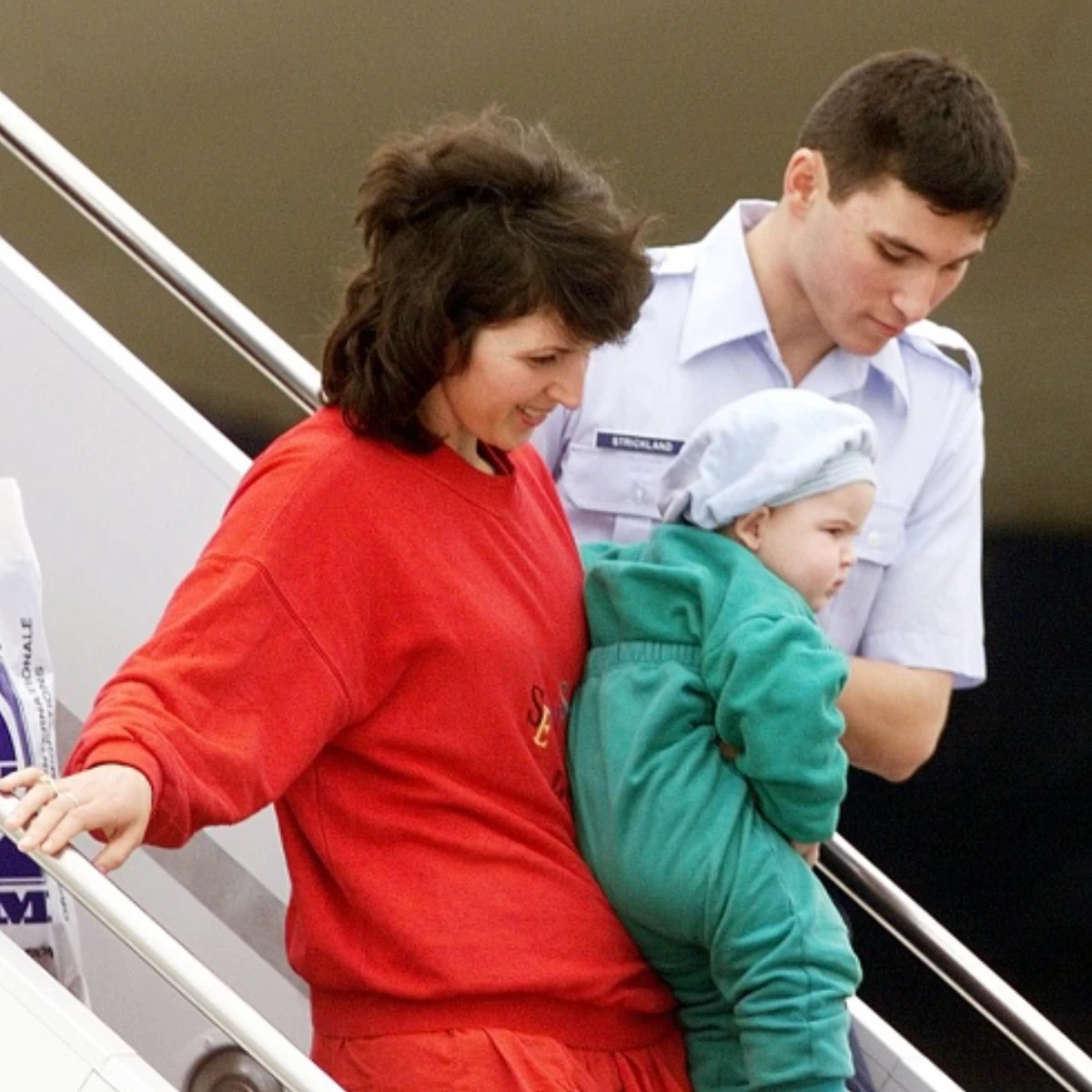 A smiling woman in a red sweatshirt carrying a baby in a green onesie. Both are being assisted down an airplane stairway by a uniformed serviceman.