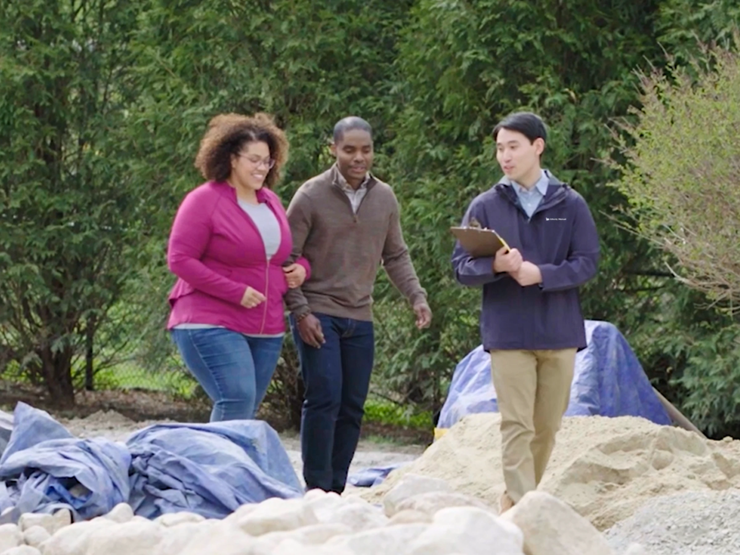 A dark-skinned man walking arm-in-arm on a sunny day with a medium-skinned woman along a rocky path with a backdrop of trees. A light-skinned person walks beside them carrying a clipboard