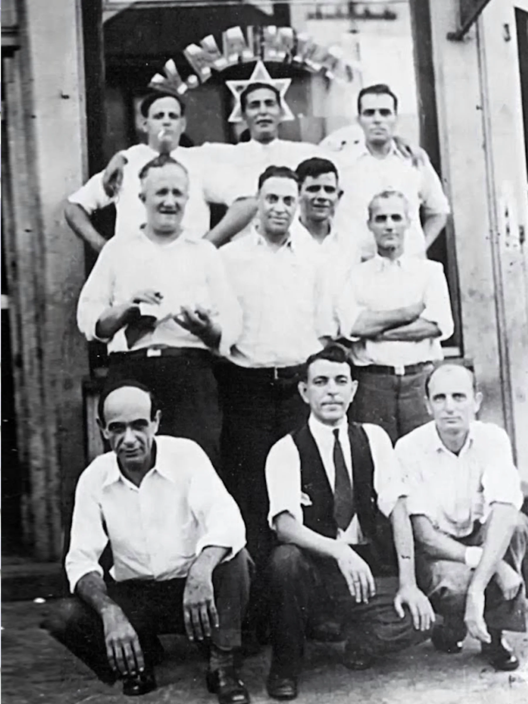 Black and white portrait of ten light-skinned men, some standing and others crouched, in front of a doorway with a sign reading "Y. Nahum" above a six-pointed star.