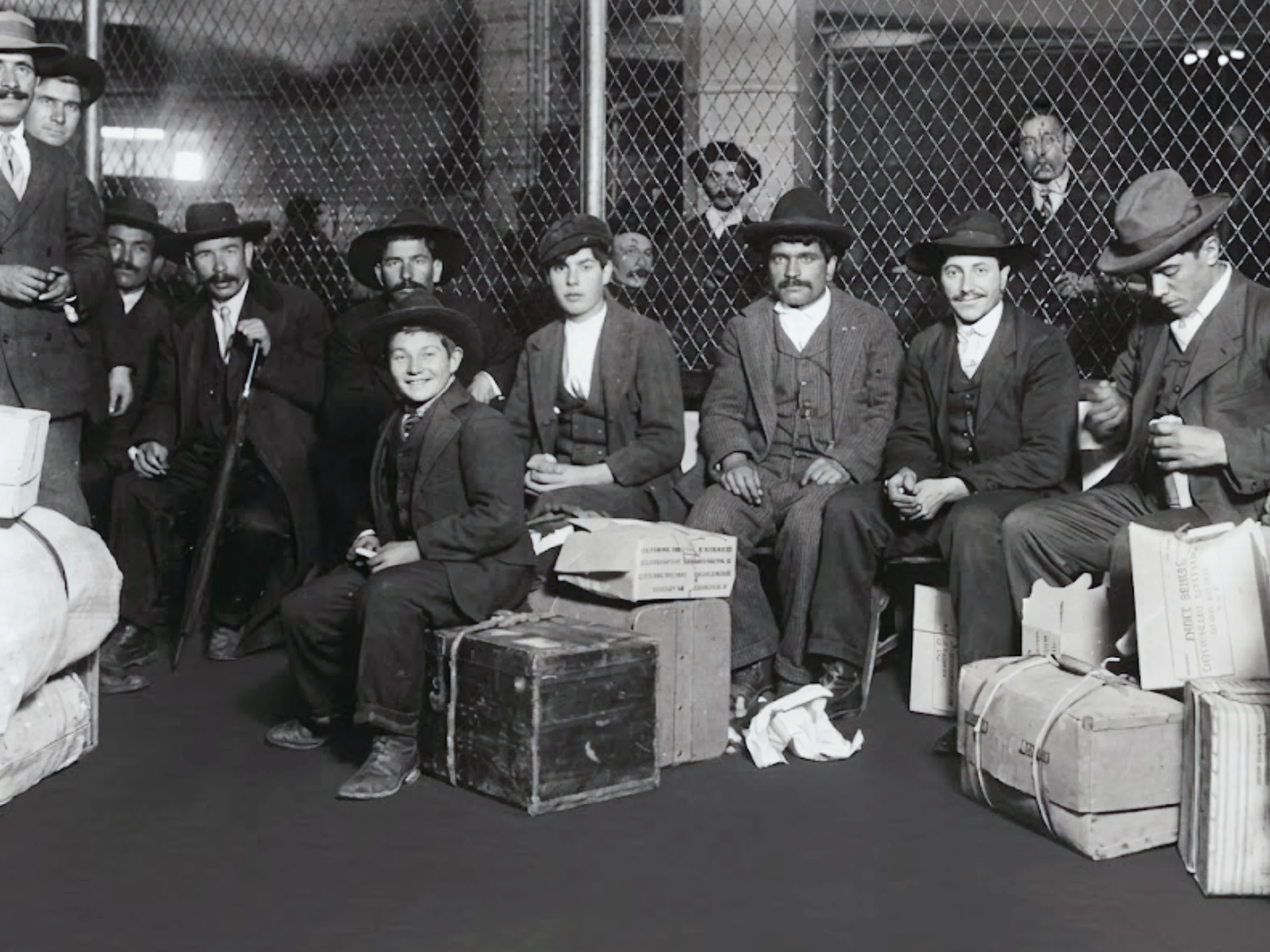 Black and white group photo of men seated with their luggage in front of a tall chain-link fence, with other people standing behind the fence.