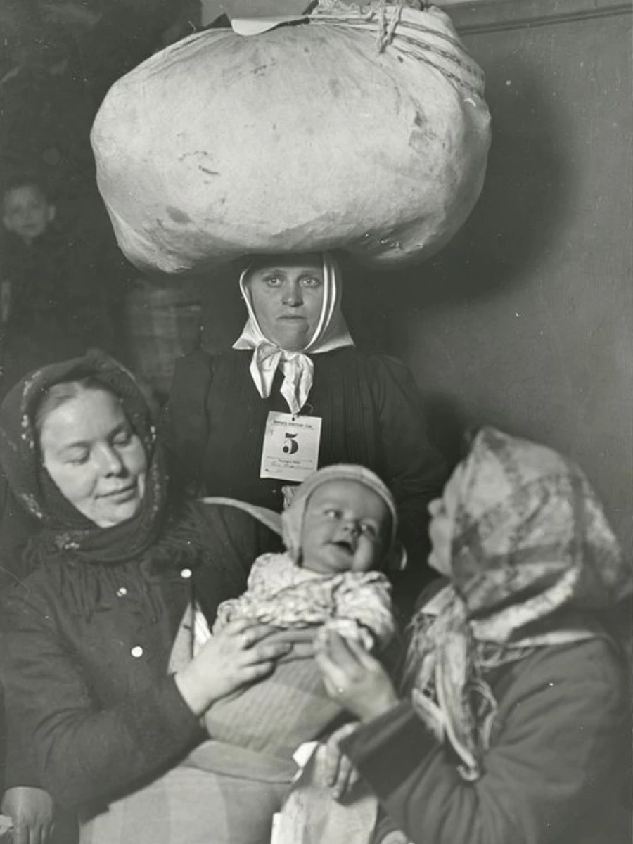 Early 20th century black and white photo of three women in head coverings gathered around a baby. Two look at the baby while a third stands behind them with a large textile sack on her head and a card printed with the number 5 pinned to her chest.