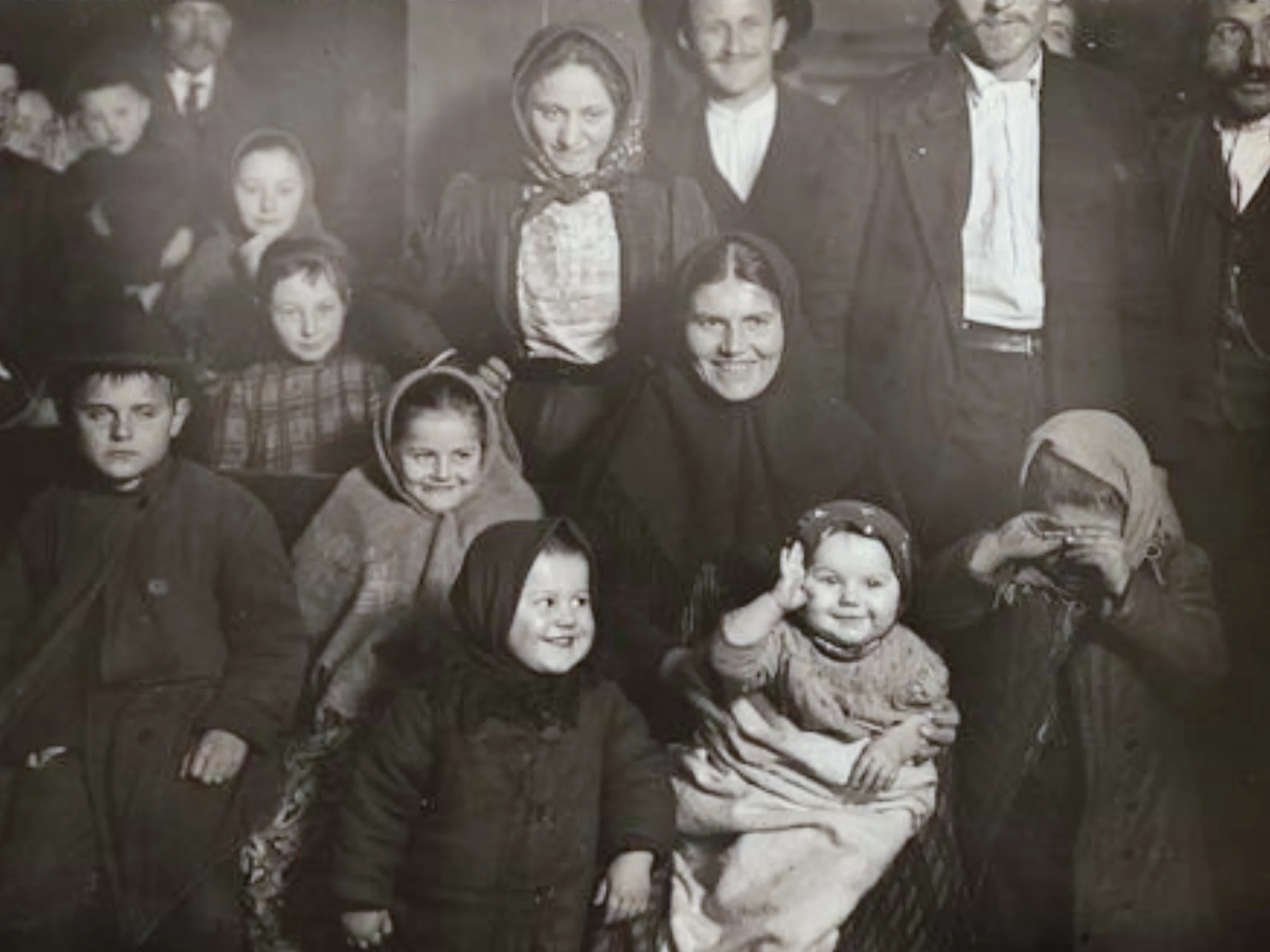 A black and white group photo of various men, women, and children posing together in a dimly lit room, wearing traditional attire and a variety of expressions.