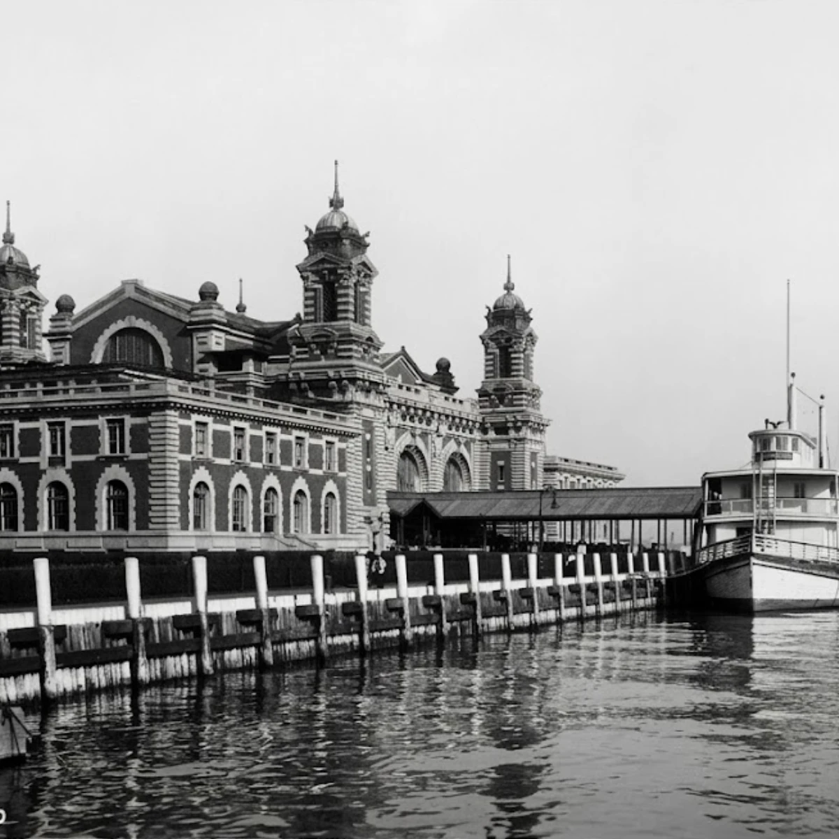 A black and white historic image of the Ellis Island Museum’s Main Building waterfront, featuring a large ornate brick building adjacent to a docked boat.