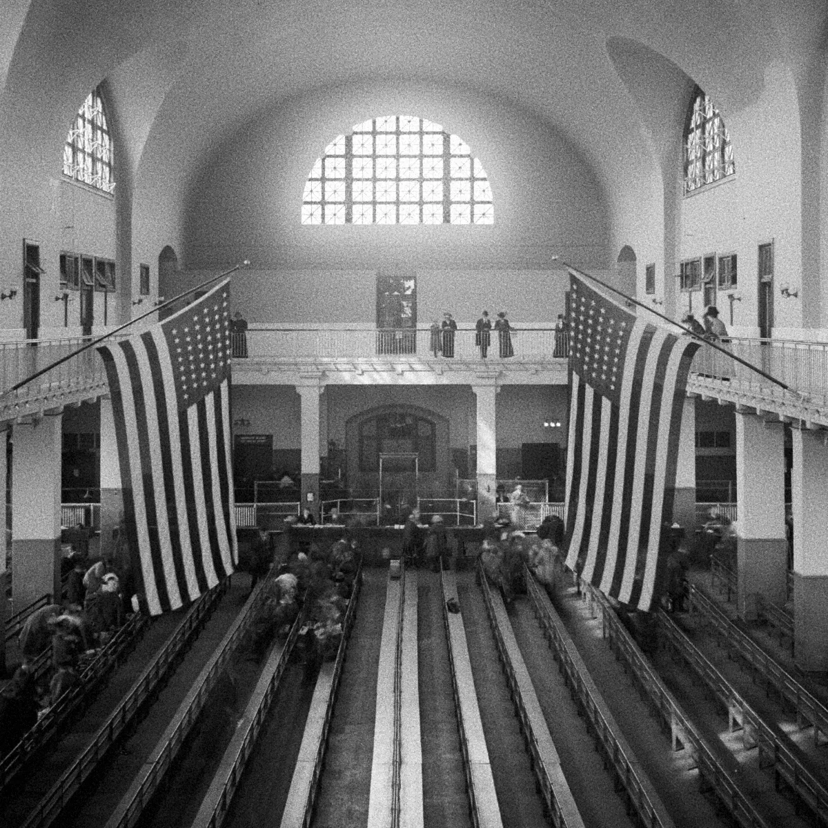 A black and white photo taken from the mezzanine of the Great Hall at Ellis Island. Two large American flags hang from either side, with rows of benches seating people below, and arched ceilings illuminated by natural light through large windows.