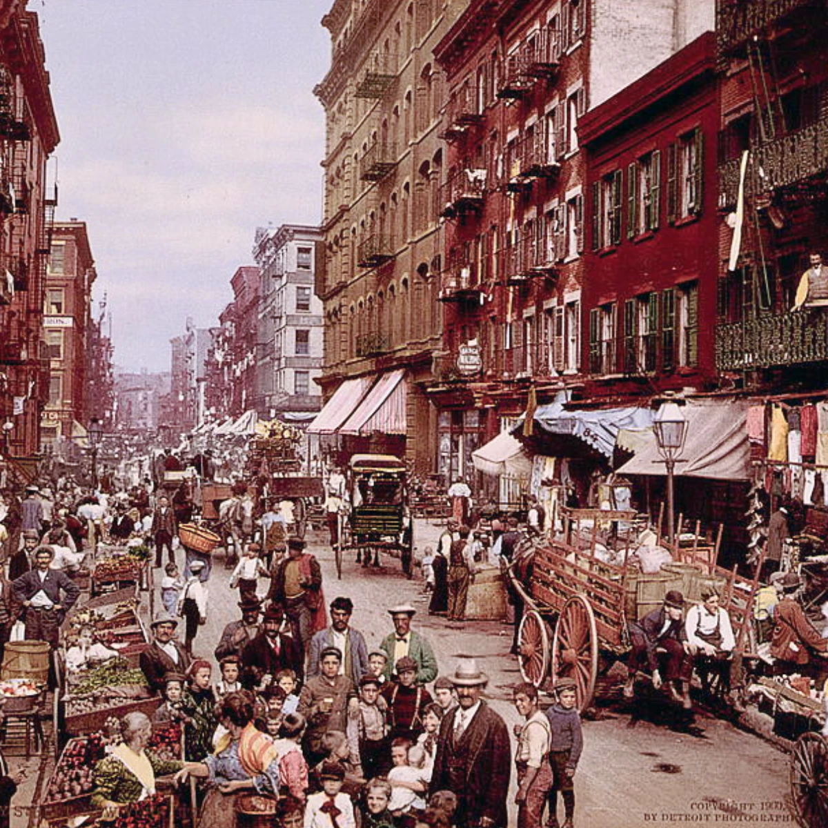A color, vintage photo of a busy Mulberry Street in early 20th-century New York City, with crowds, market stalls, and horse-drawn carriages. Various men, women, and children in period attire fill the foreground.