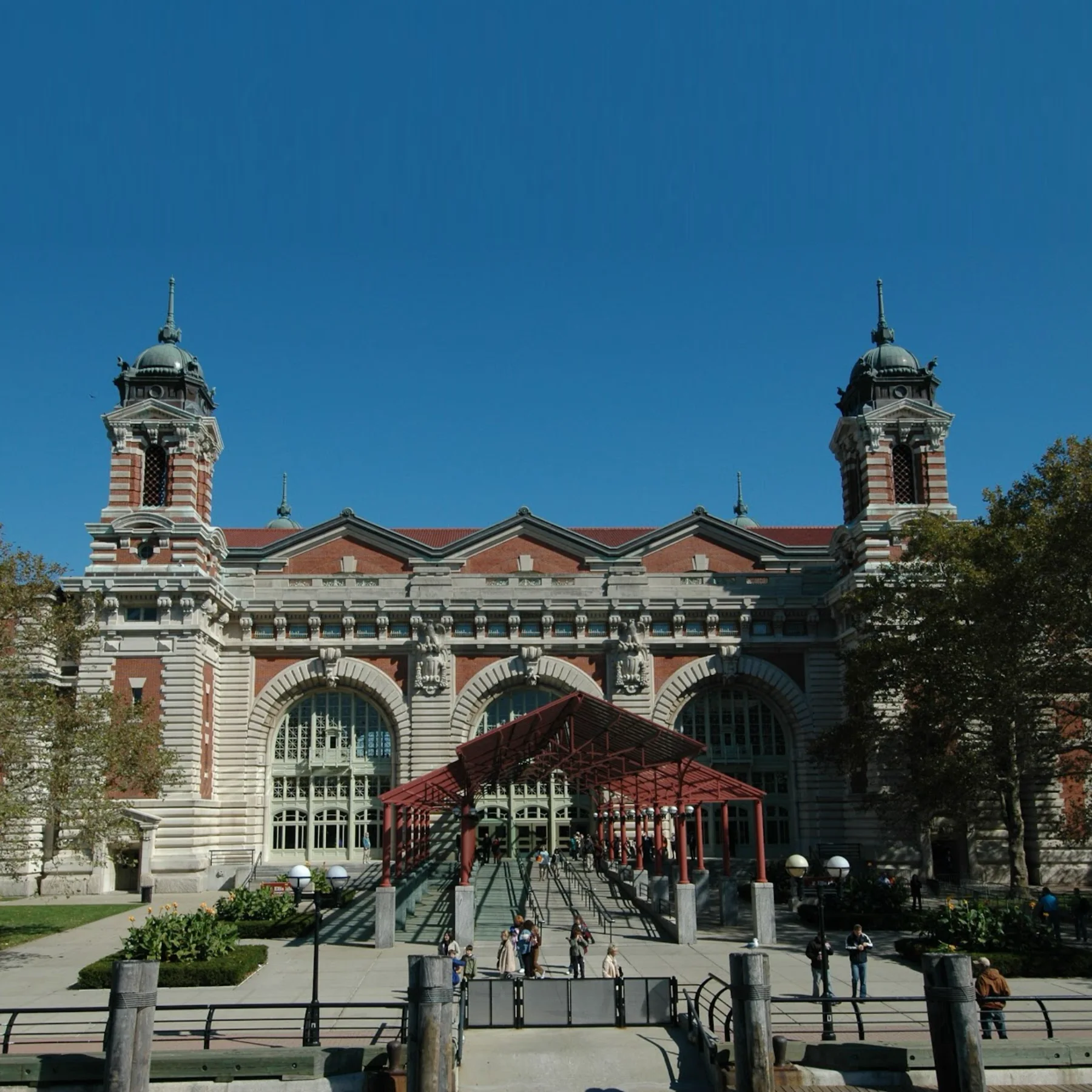 The front facade of the Ellis Island main building. Two towers with green copper-domed tops and spires rise from the red brick building. A red metal canopy structure covers the walkway leading to the entrance.