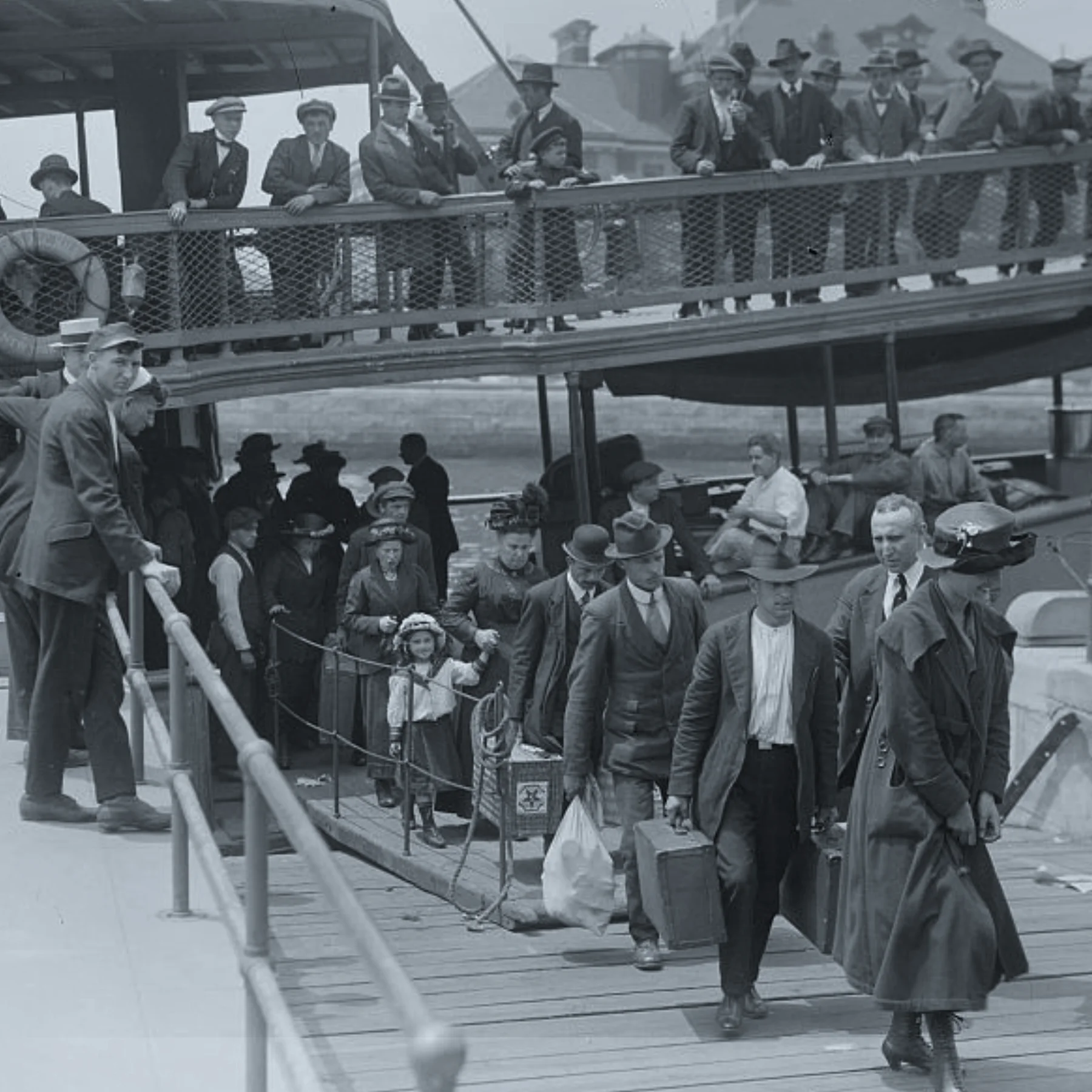 Black and white photo of a long line of people disembarking a boat down a ramp above, and walking toward the camera up a wooden gangplank below. They carry luggage, and cloth bundles.