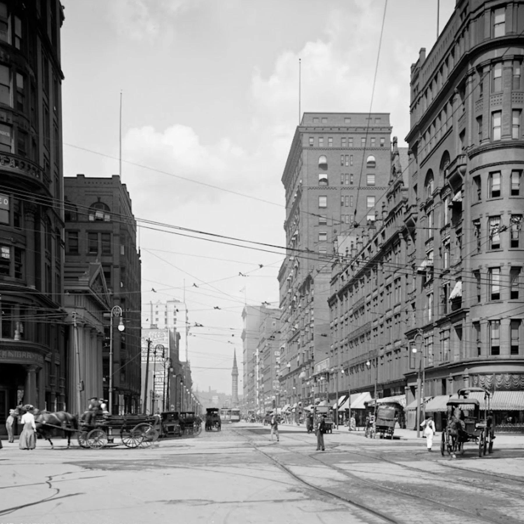 Black and white early 20th-century city street featuring historic stone buildings, horse-drawn carriages, streetcar tracks, dense overhead wires, and pedestrians on sidewalks.