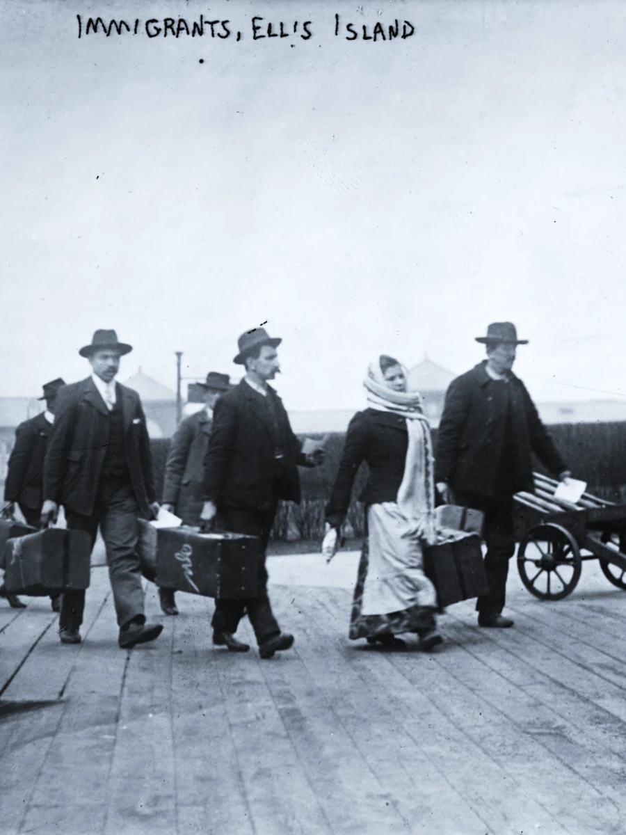 A black and white photo of five people walking on the wooden boardwalk of Ellis Island, mostly dressed in dark early 20th-century clothing and carrying suitcases. A handwritten caption at the top reads "IMMIGRANTS, ELLIS ISLAND."