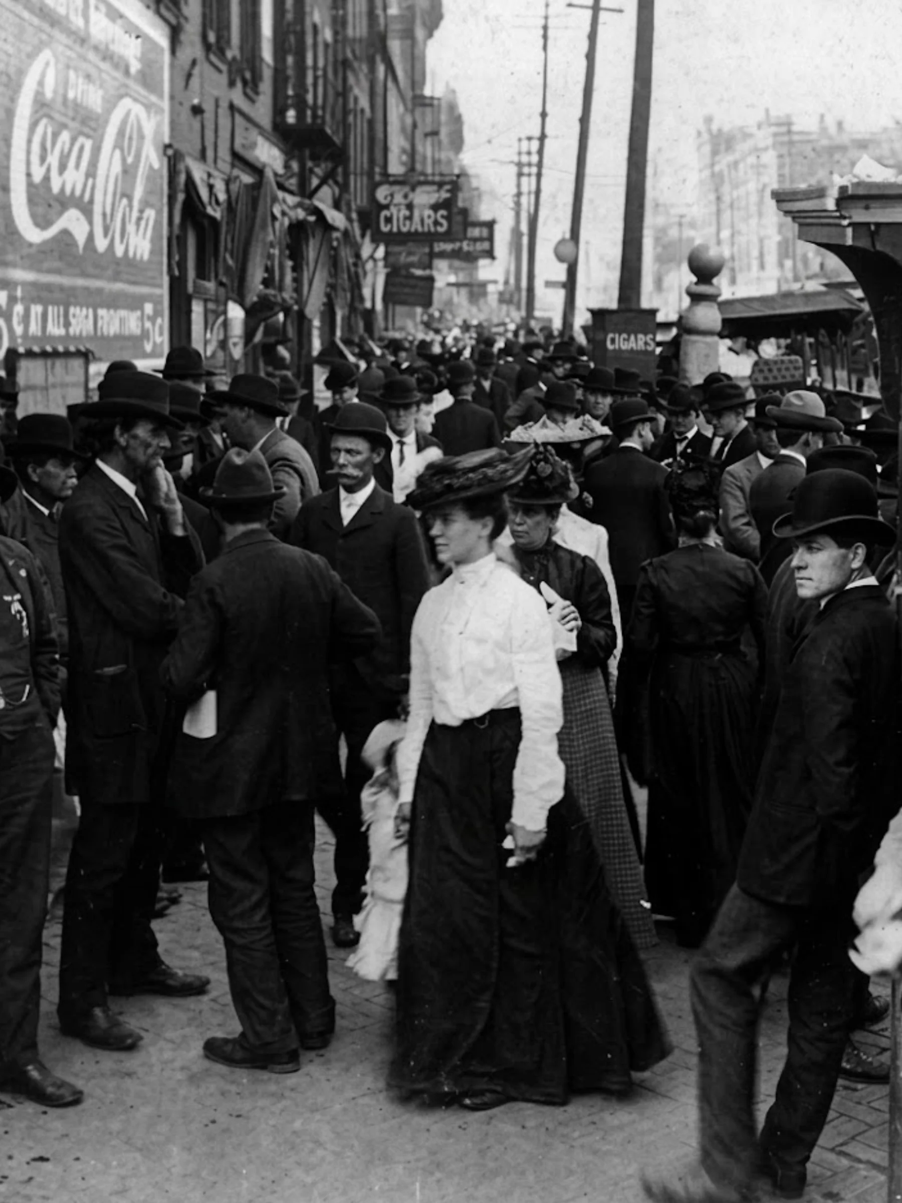 Black and white early 20th-century photo of a crowded street filled with men in bowler hats and women in elaborate headwear, walking past vintage Coca-Cola and cigar signs.