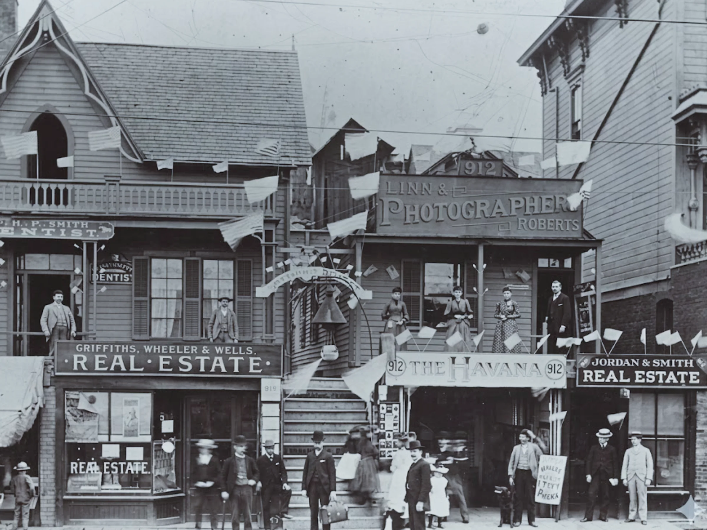 Black-and-white, late 1800s photo of two-story wooden storefronts with real estate and photography signs, decorated with flags, and people on balconies and sidewalks.